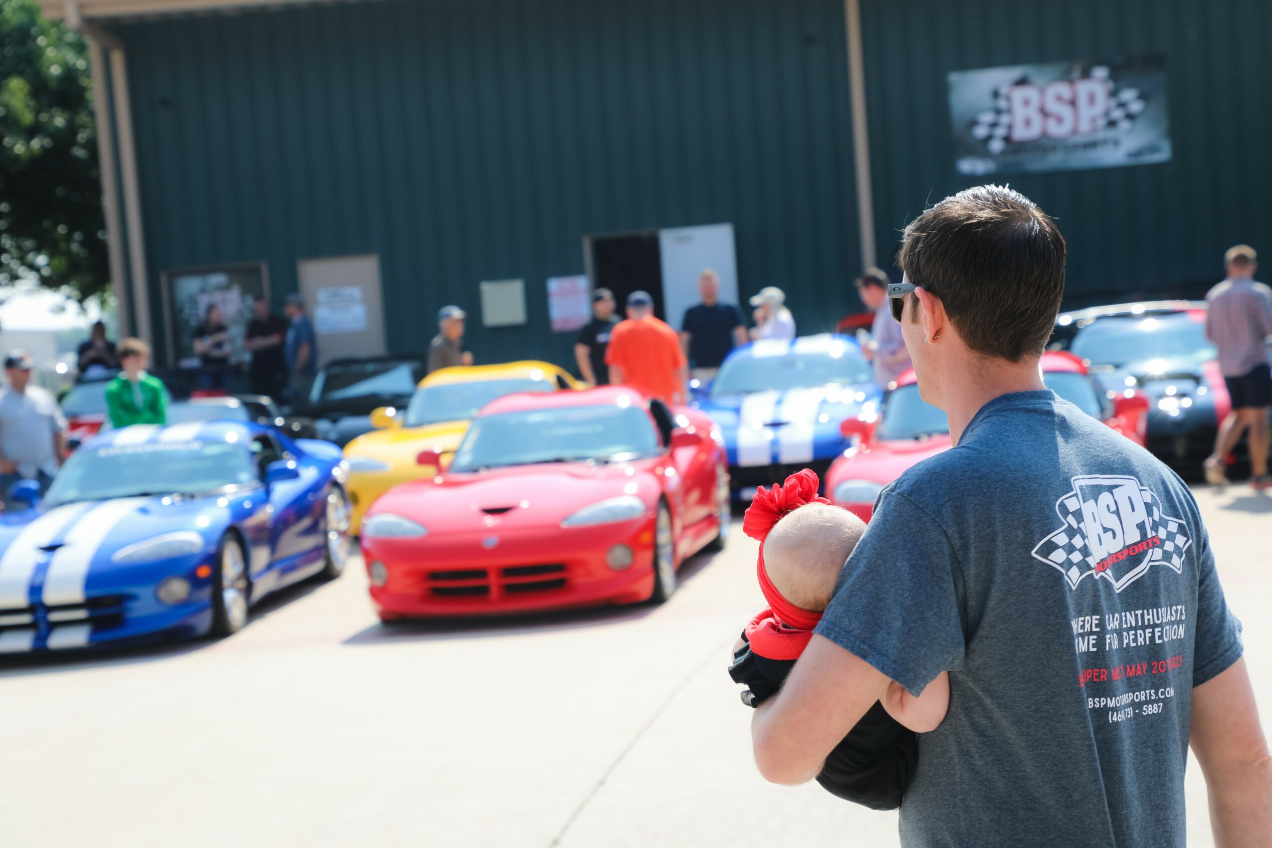 Man holding a child at a car show with colorful sports cars parked outdoors.