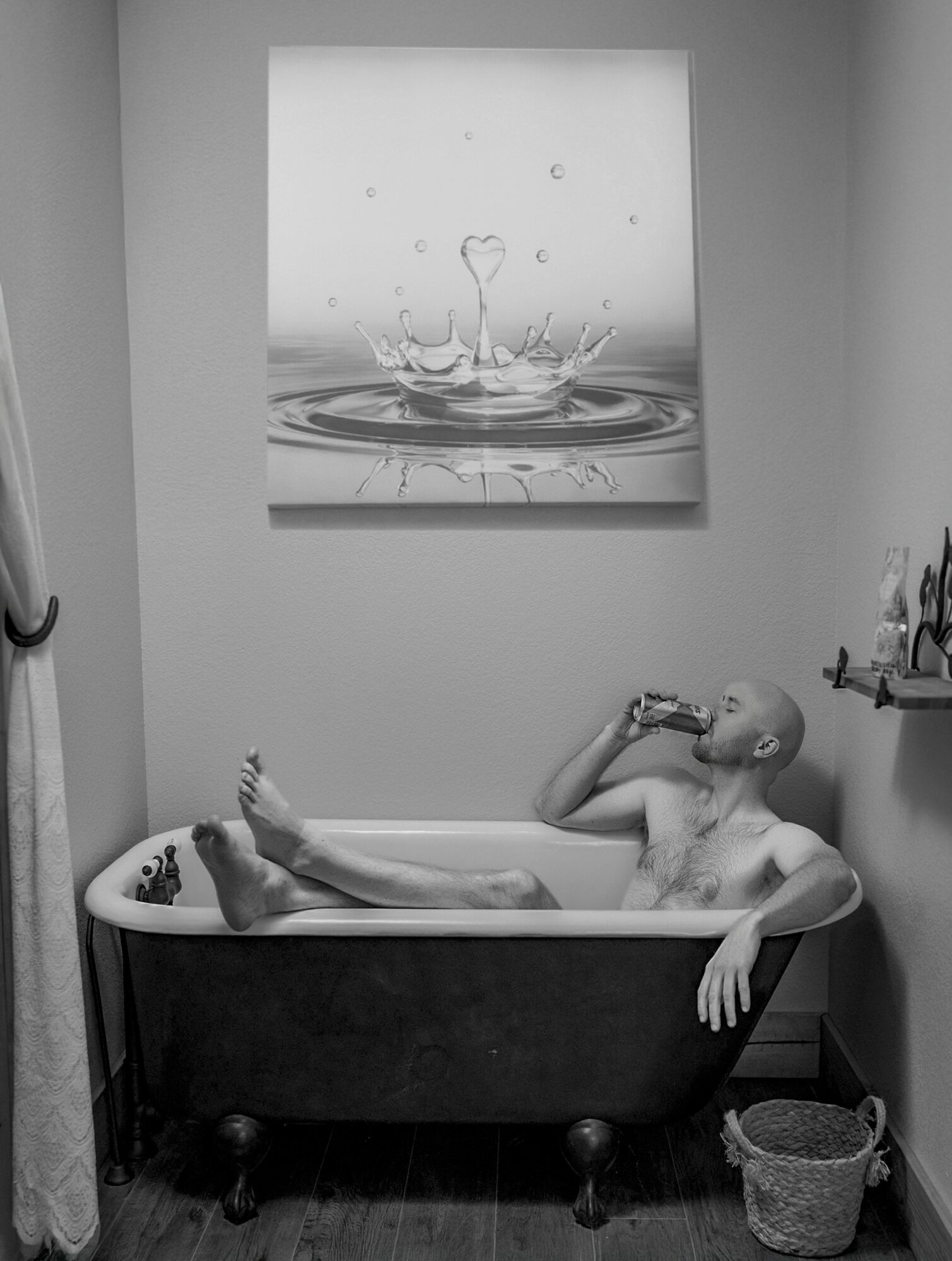 Person with shaved head relaxing in a bathtub, drinking from a glass, with a painting of a water droplet above.
