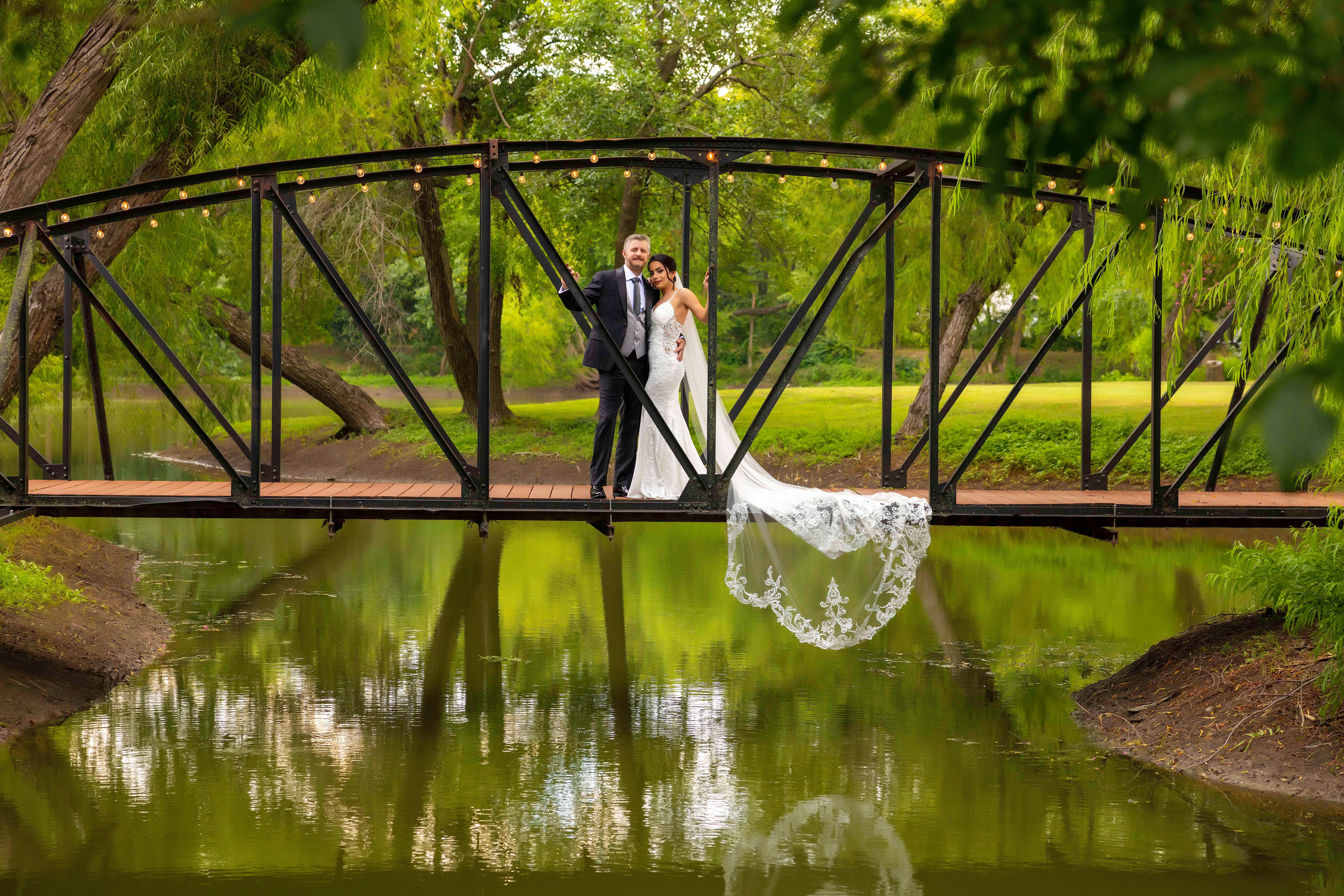 Bride and groom standing on a bridge over a pond, surrounded by green trees, with the bride's dress hanging over the edge.