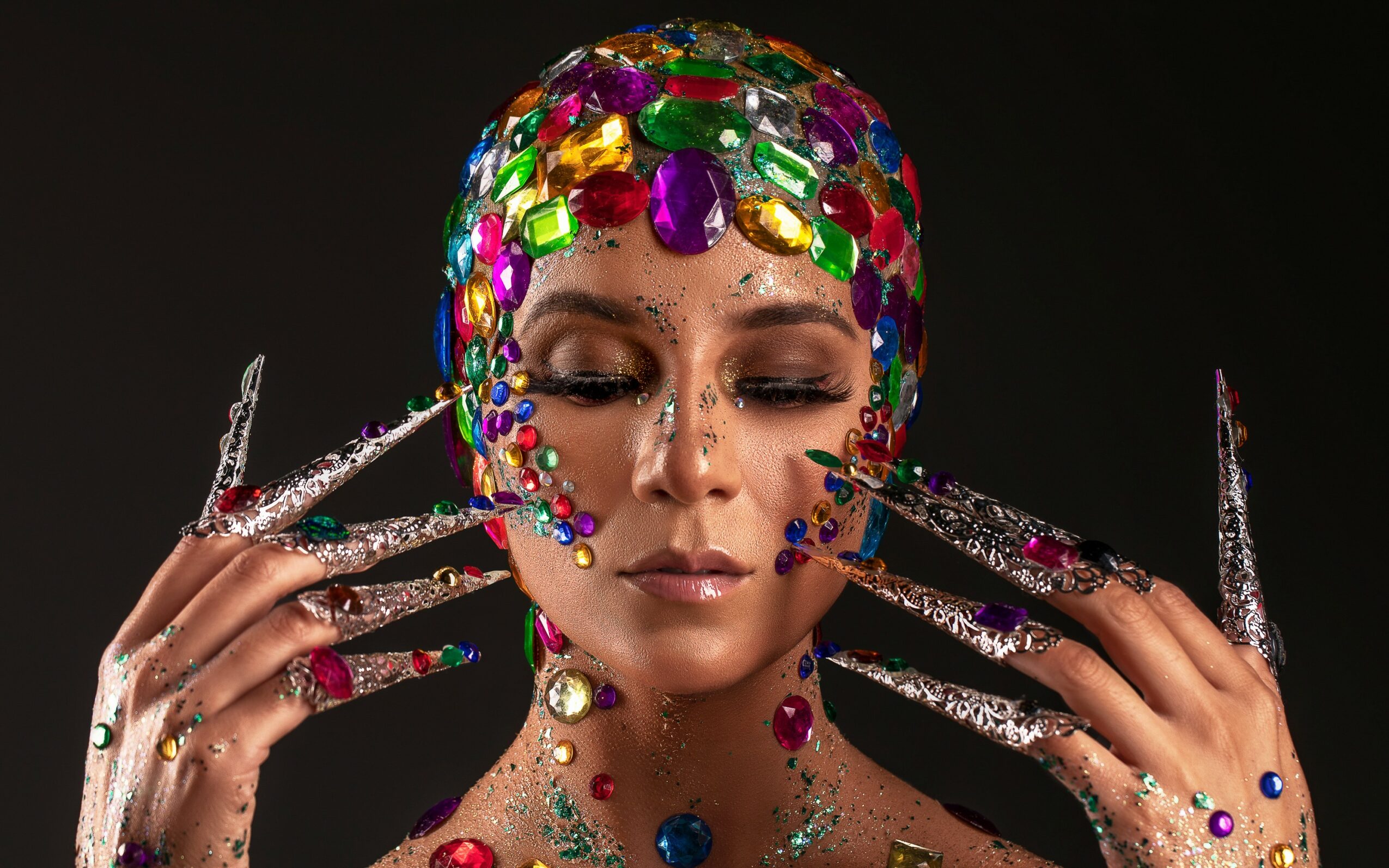 Woman with colorful gemstone headpiece and matching jewelry, holding her face with long, decorated nails.
