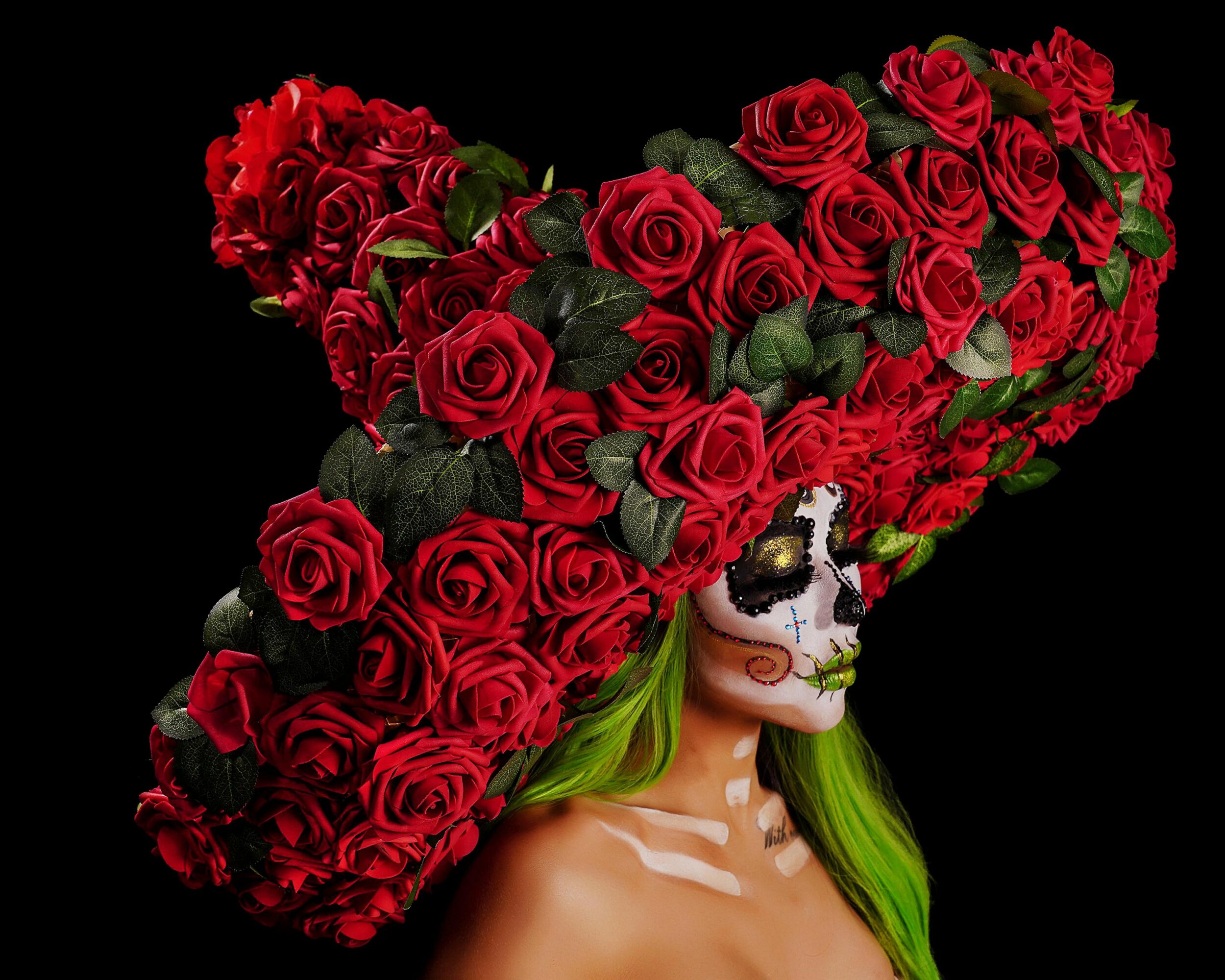 Person with face painted as a skull, green hair, and a large hat made of red roses against a black background.