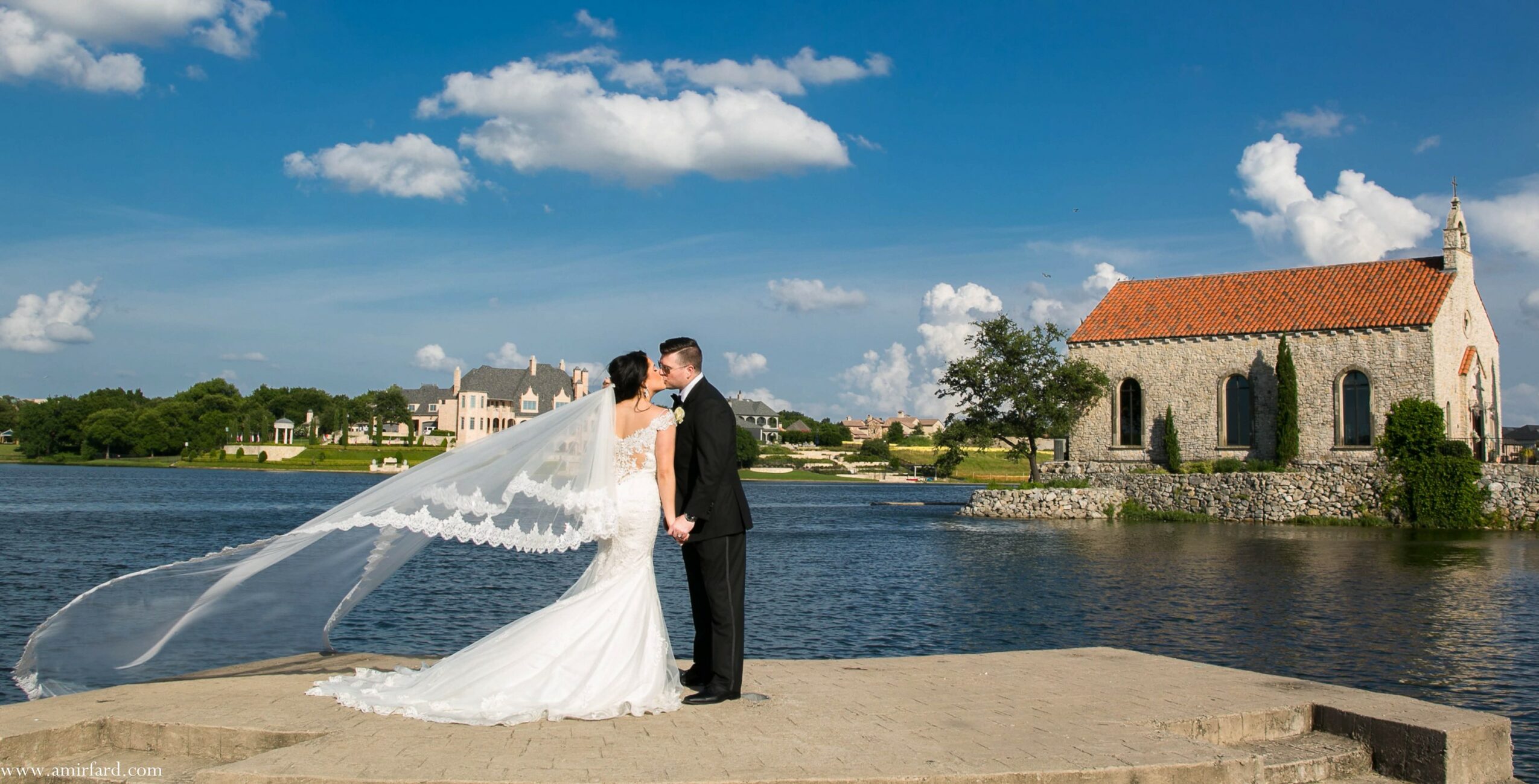 Bride and groom standing on a beach near water, with a church and trees in the background under a blue sky.
