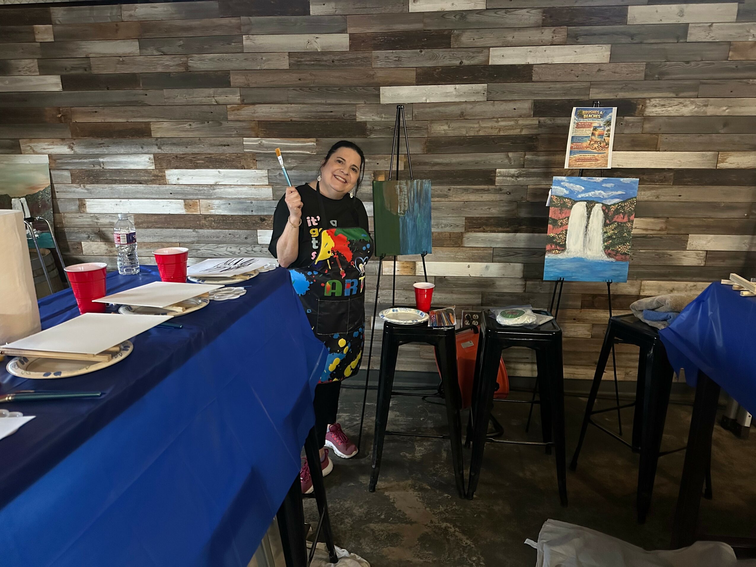 Person standing next to an easel with a painting, holding a paintbrush, in a room with wooden wall and tables.