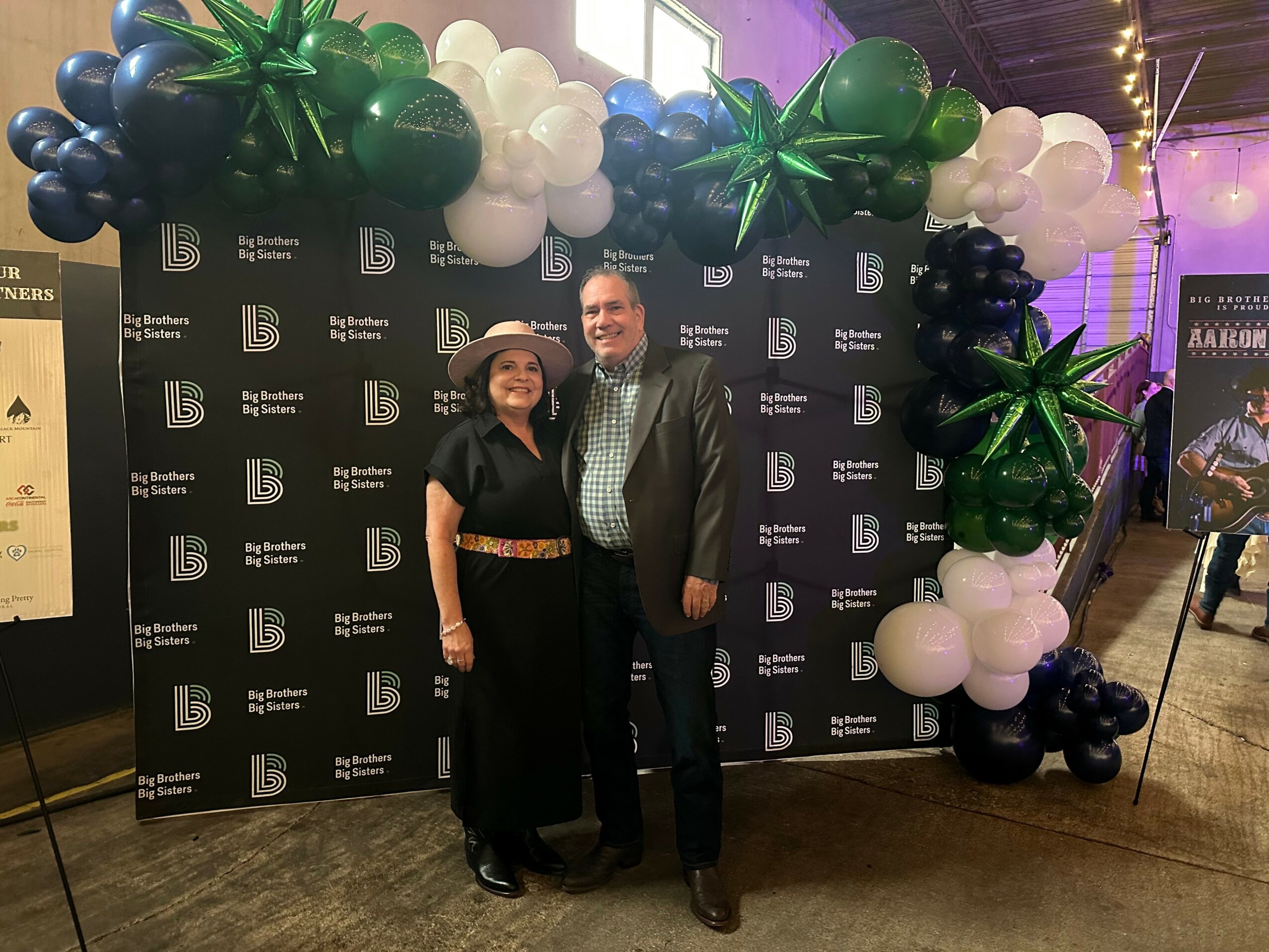 Two people standing in front of a black backdrop with white logos, surrounded by balloon decorations, smiling at the camera.