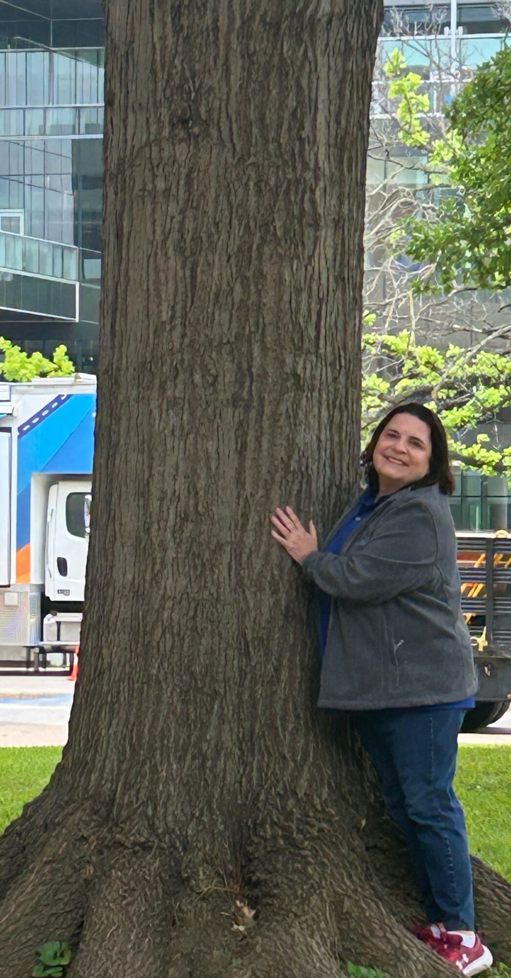 Woman hugging a large tree outdoors in an urban setting with buildings and greenery in the background.