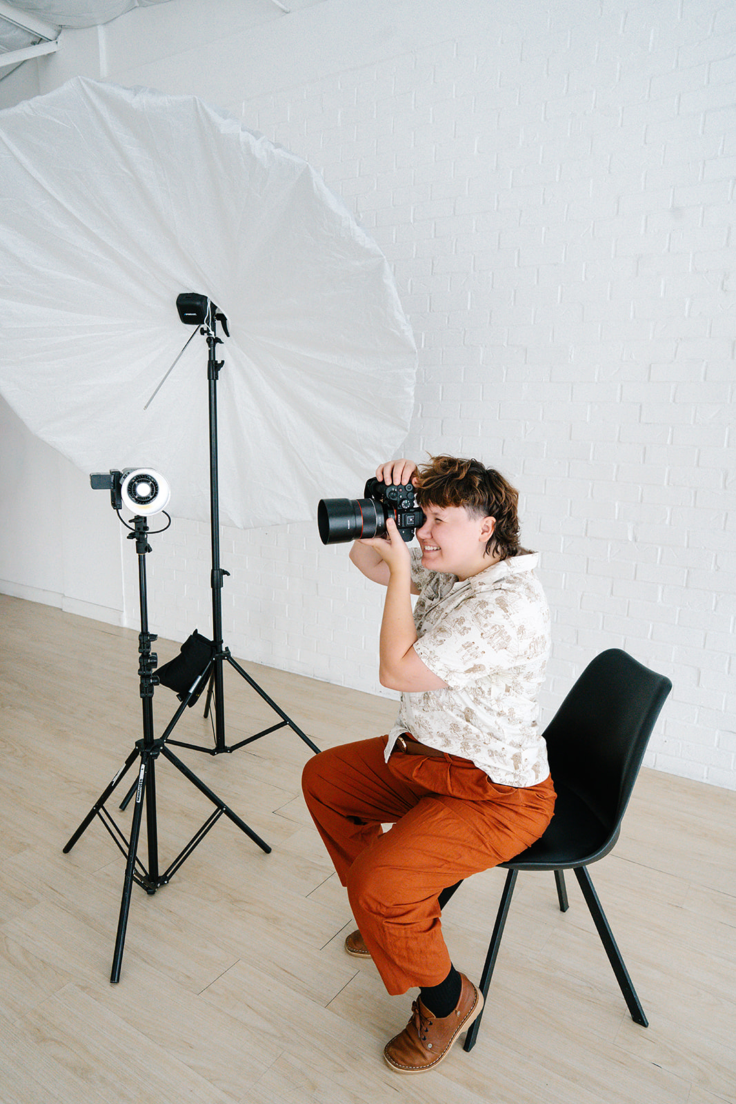 Child sitting on a black chair, holding a camera, in a photography studio with lighting equipment and a large white umbrella.