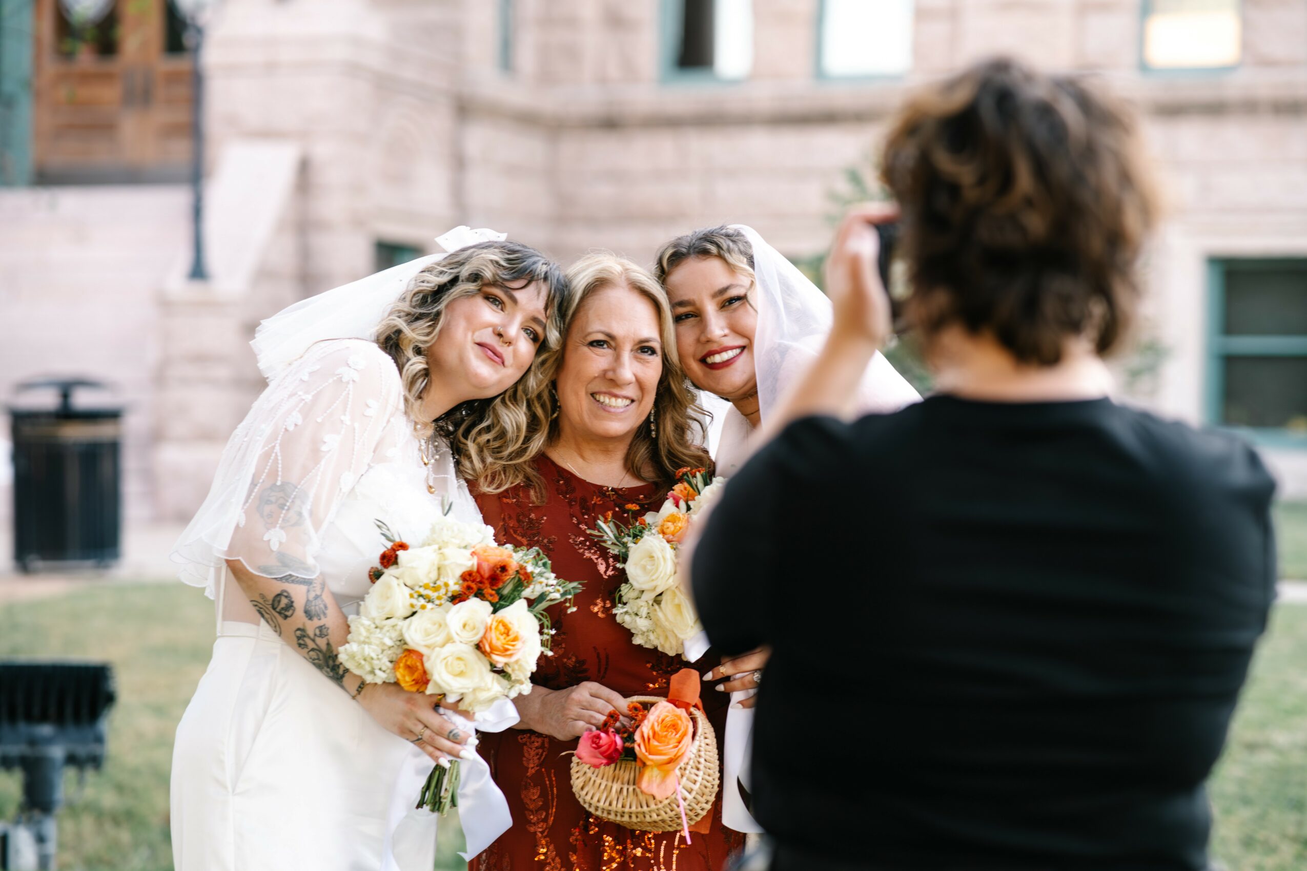 Three women smiling and holding bouquets, posing outdoors in front of a building, with a person taking their photo.