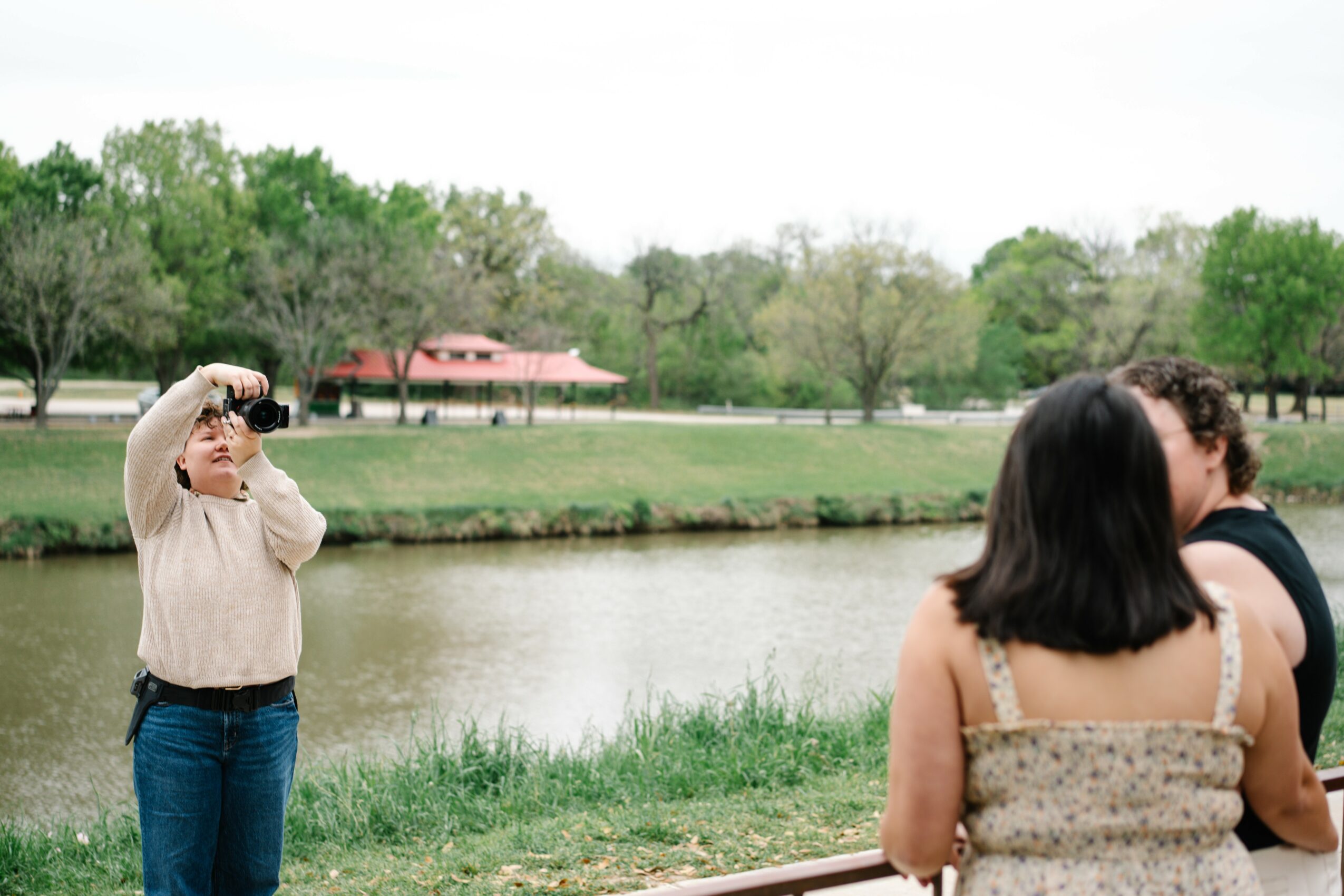 Person taking a photo of a couple near a river with trees and a pavilion in the background.