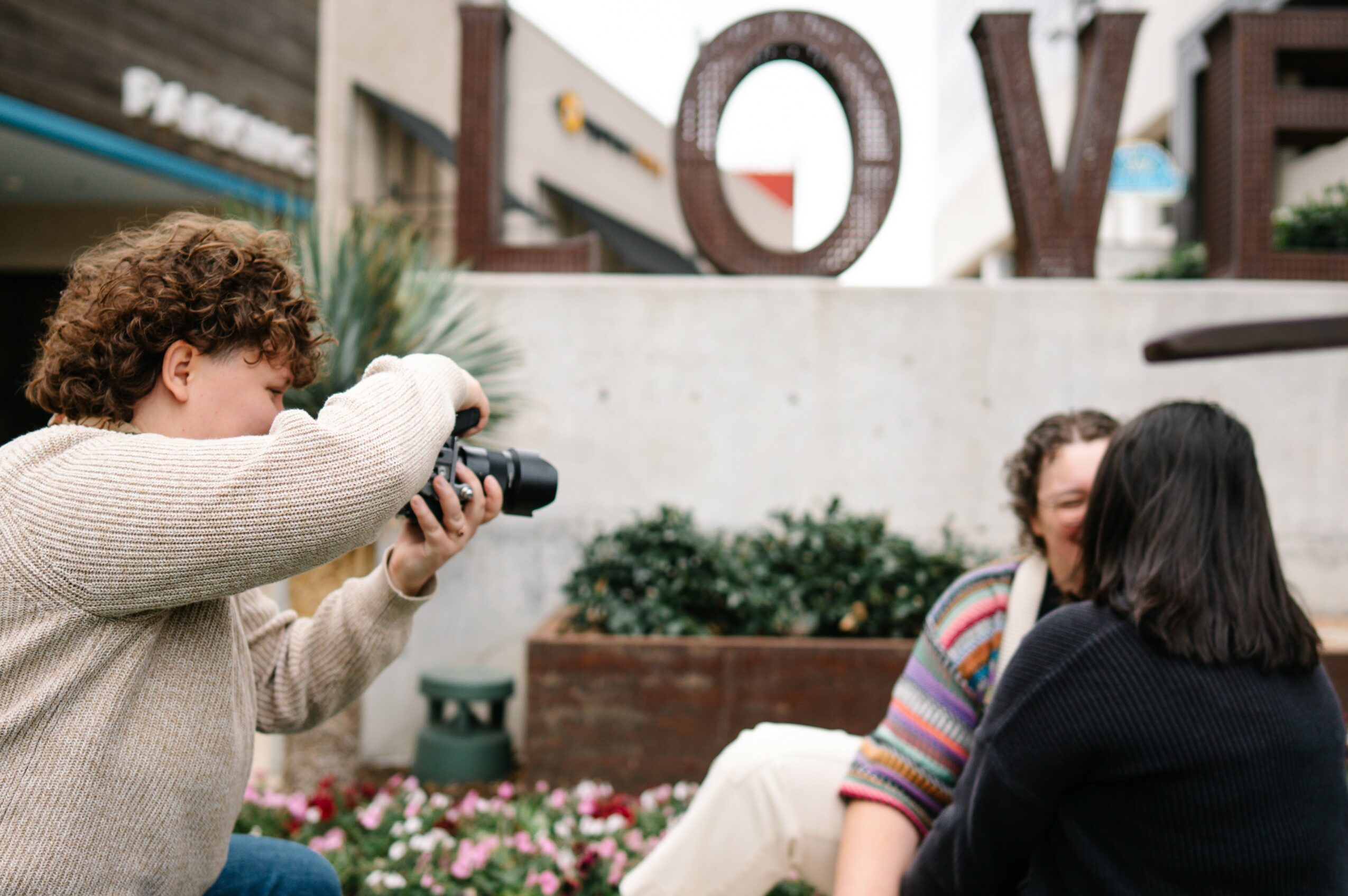 Two women sitting outdoors, one taking a photo of the other, with large letters spelling 'LOVE' in the background.