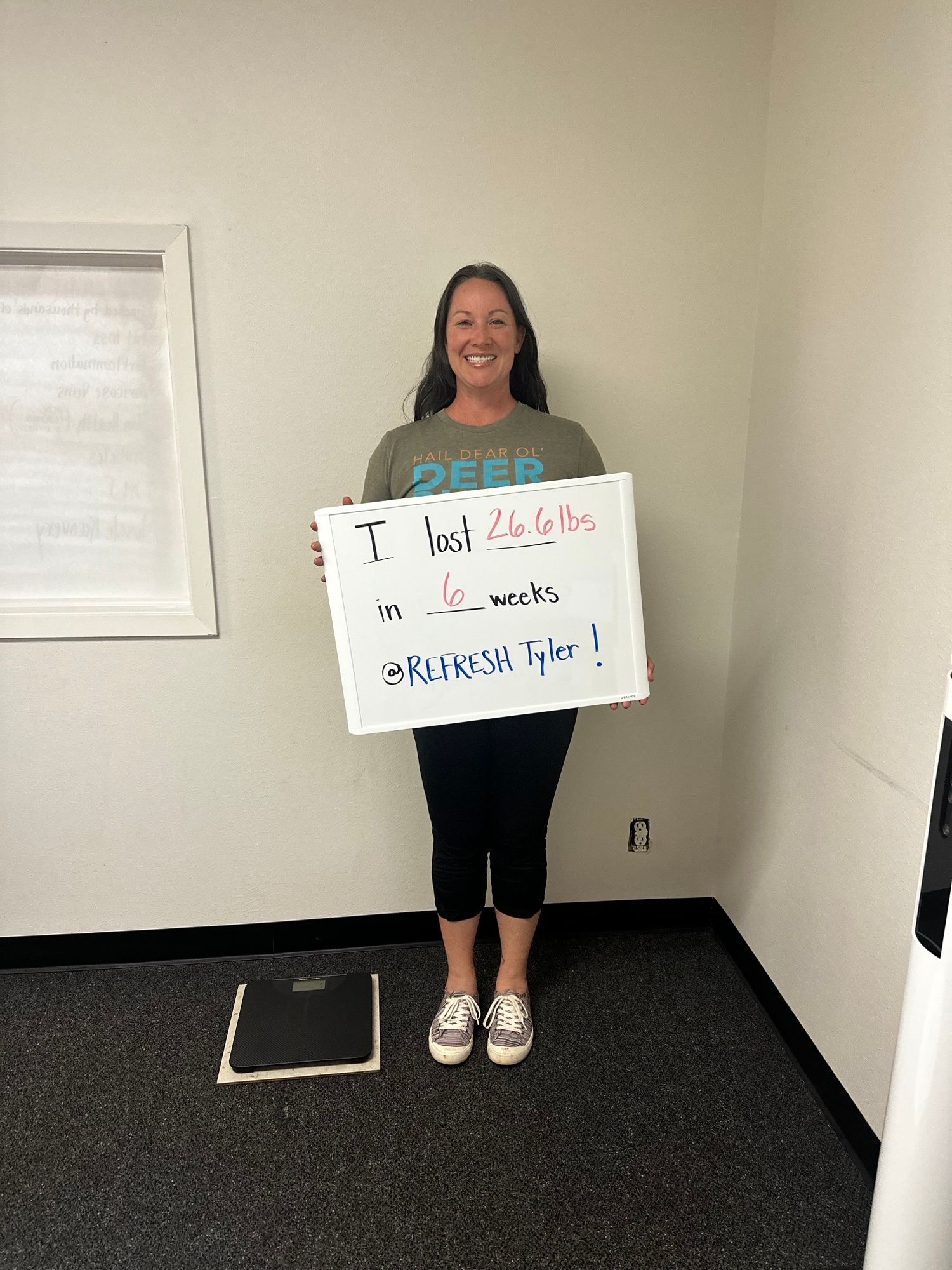 Woman smiling holding a sign with handwritten message in a room with white walls and black carpet.