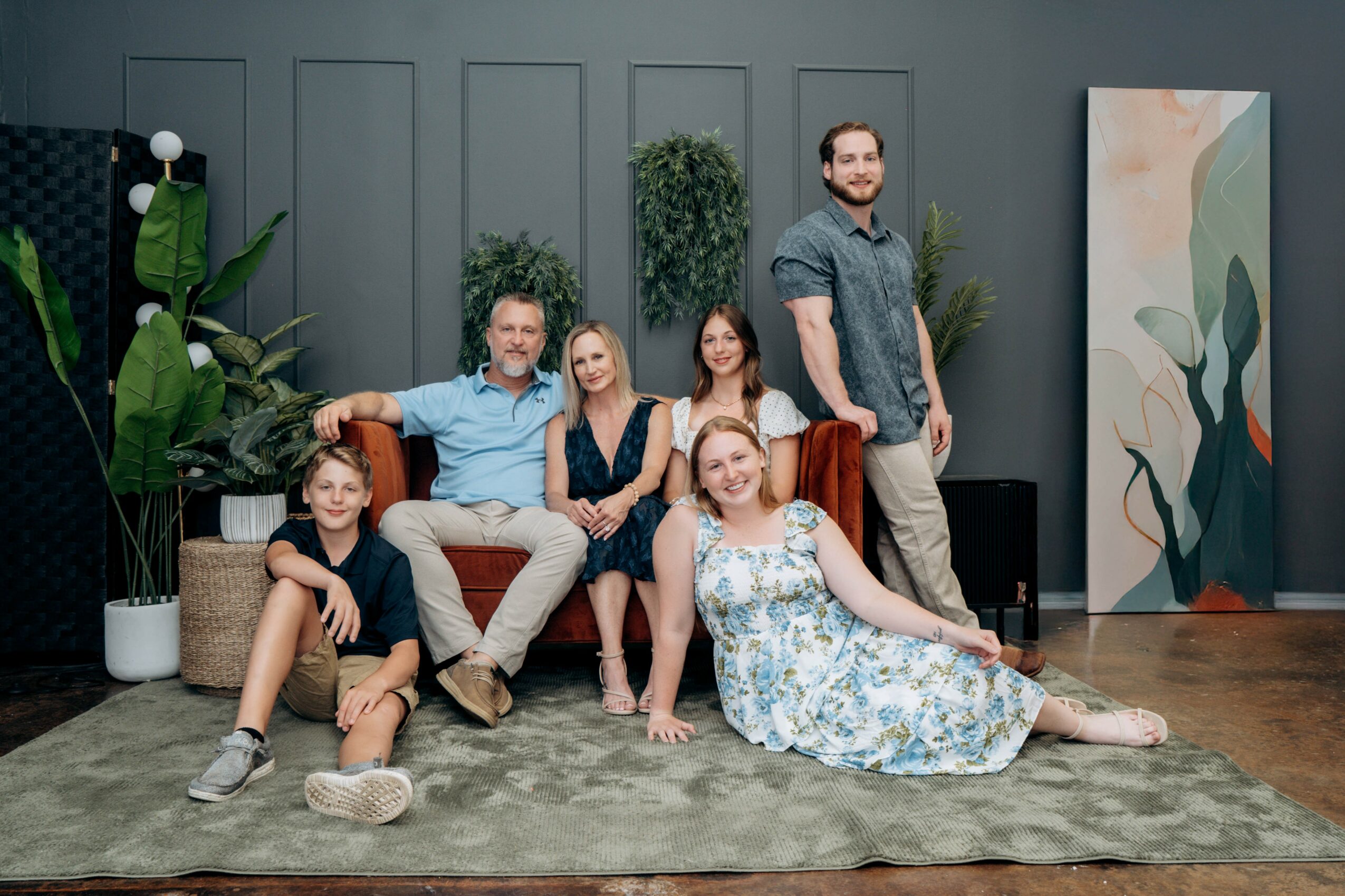 Group of seven people sitting and lying on a rug in a room with plants and artwork.