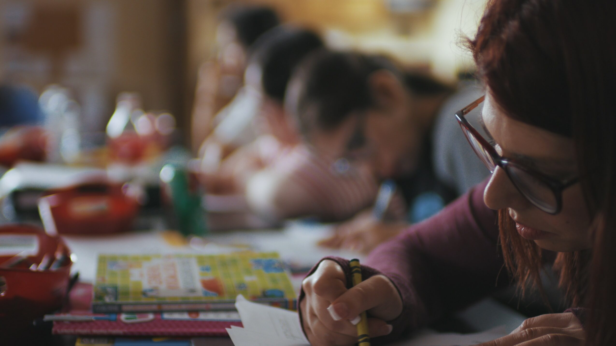 People sitting at a table, writing or drawing, with colorful papers and supplies, in a classroom or workshop setting.