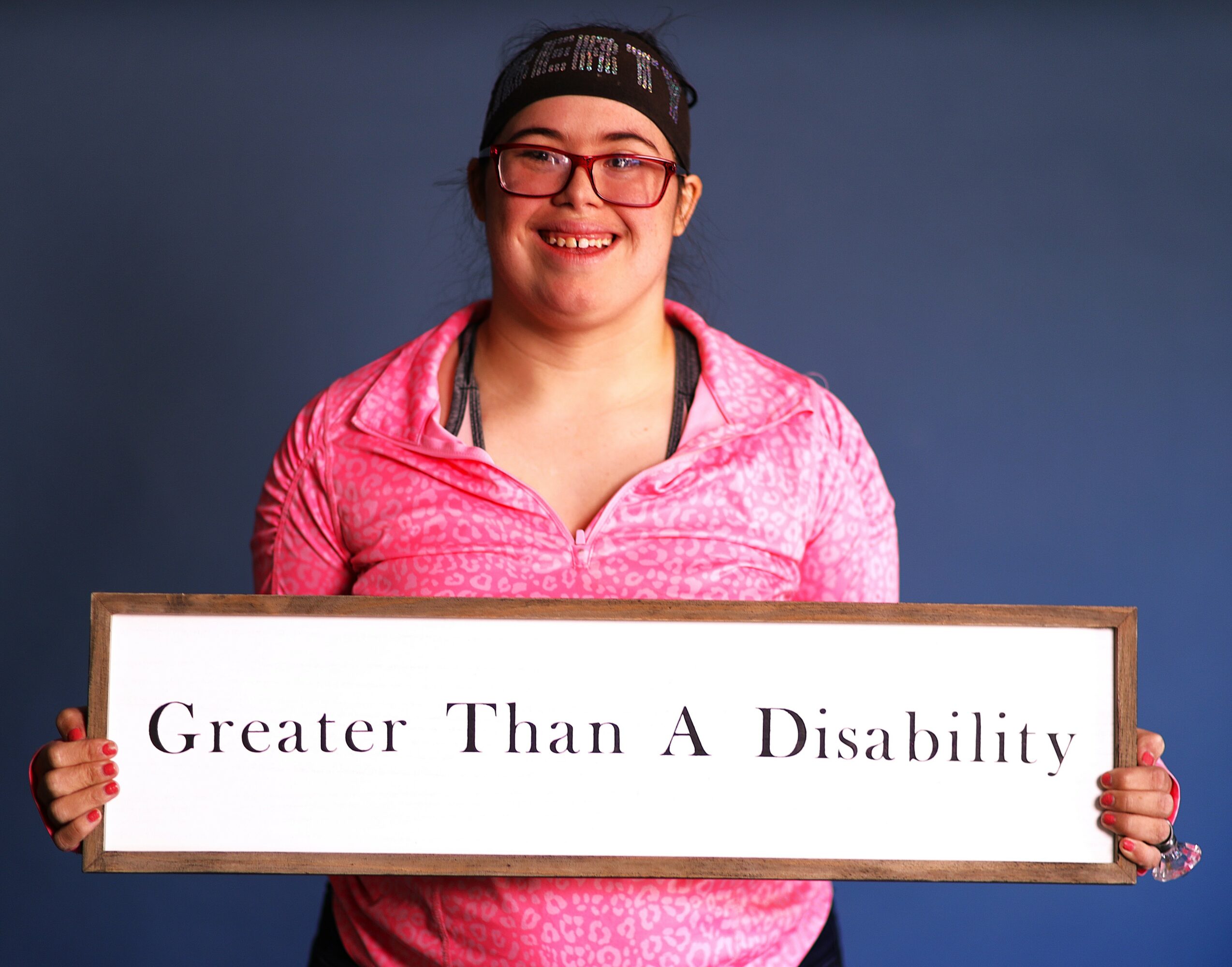 Smiling woman holding a sign that reads 'Greater Than A Disability' against a blue background.
