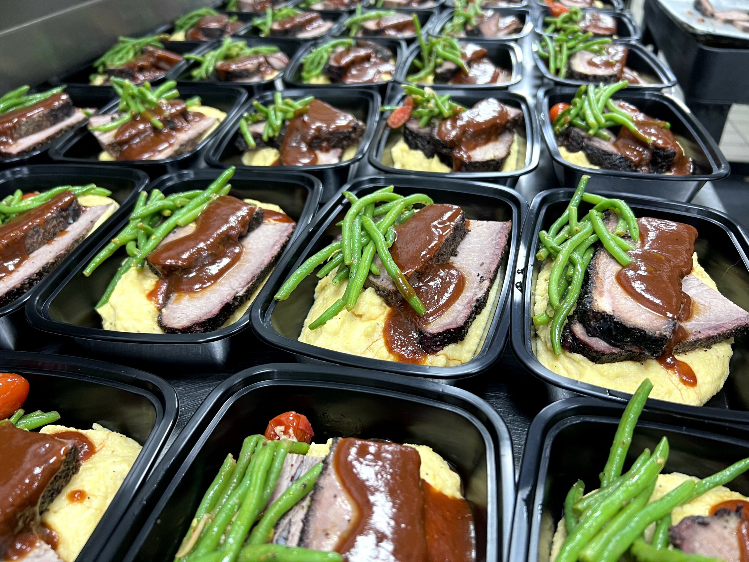 Multiple black containers with slices of meat, green beans, and mashed potatoes arranged in rows.