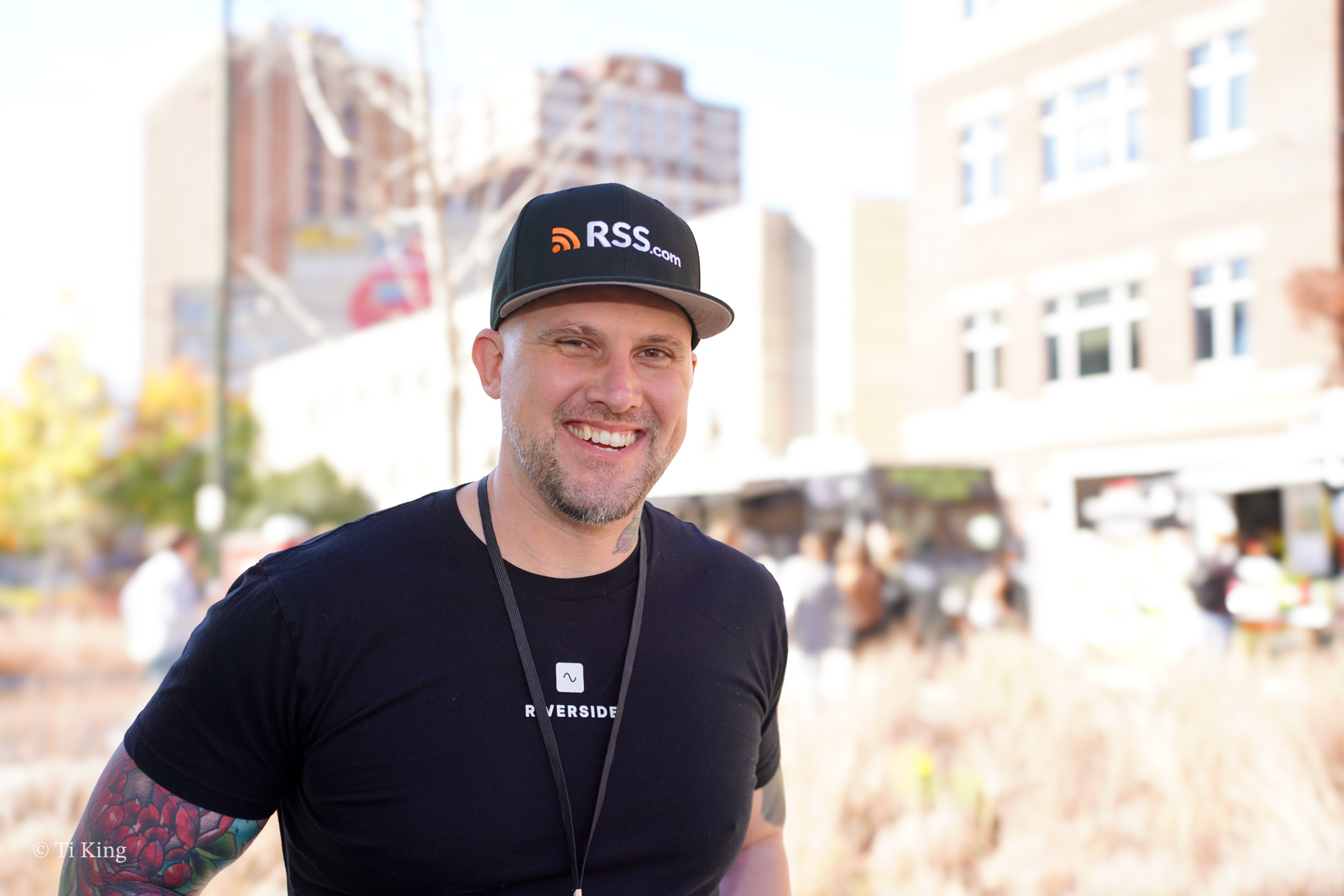 Smiling man wearing a black cap and t-shirt outdoors in an urban setting.