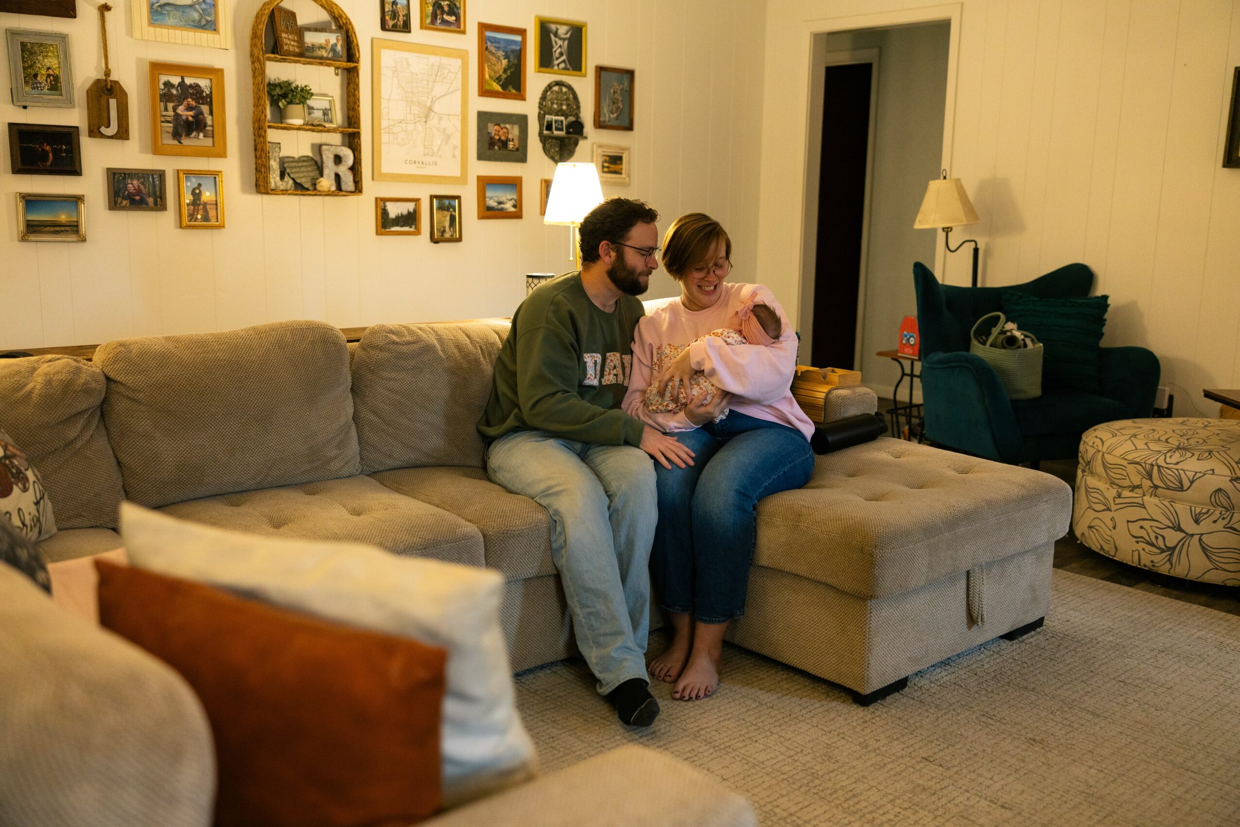 Two people sit on a beige sectional sofa in a living room, looking at a phone together. Wall with pictures behind them.