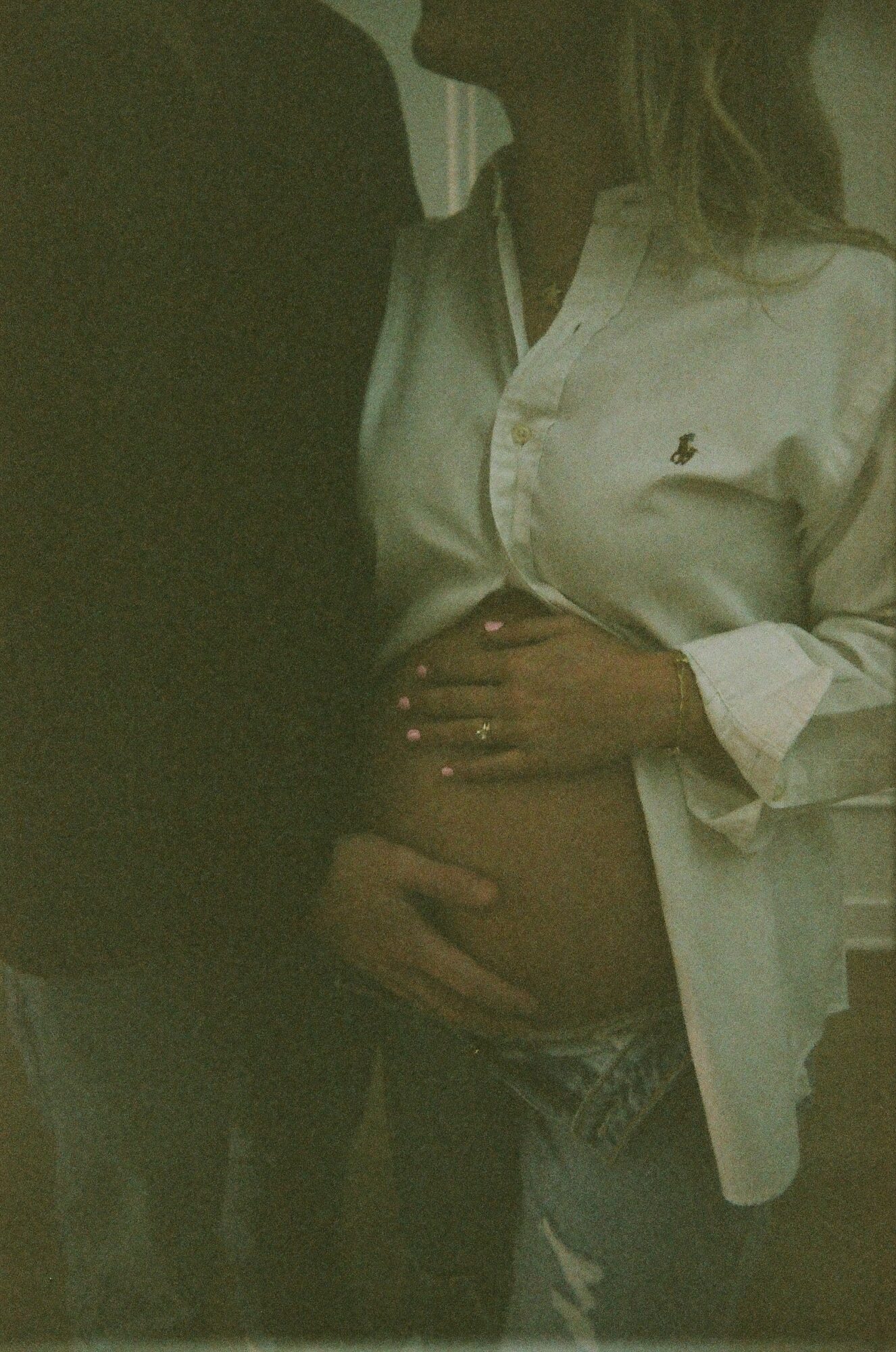 Person wearing a white shirt with a small logo, holding their pregnant belly, with another person partially visible beside them.