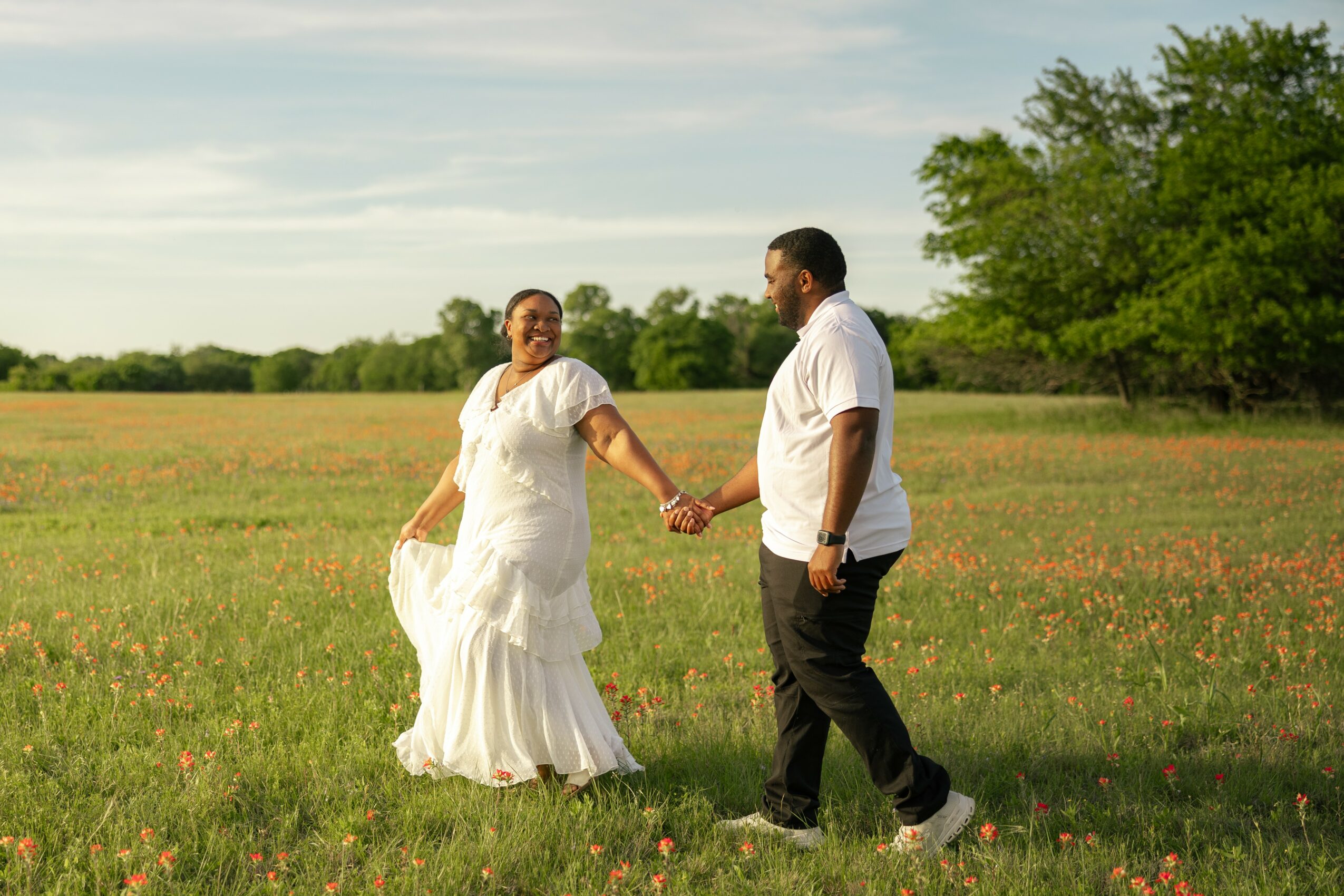 A couple holding hands and smiling in a grassy field with trees in the background.
