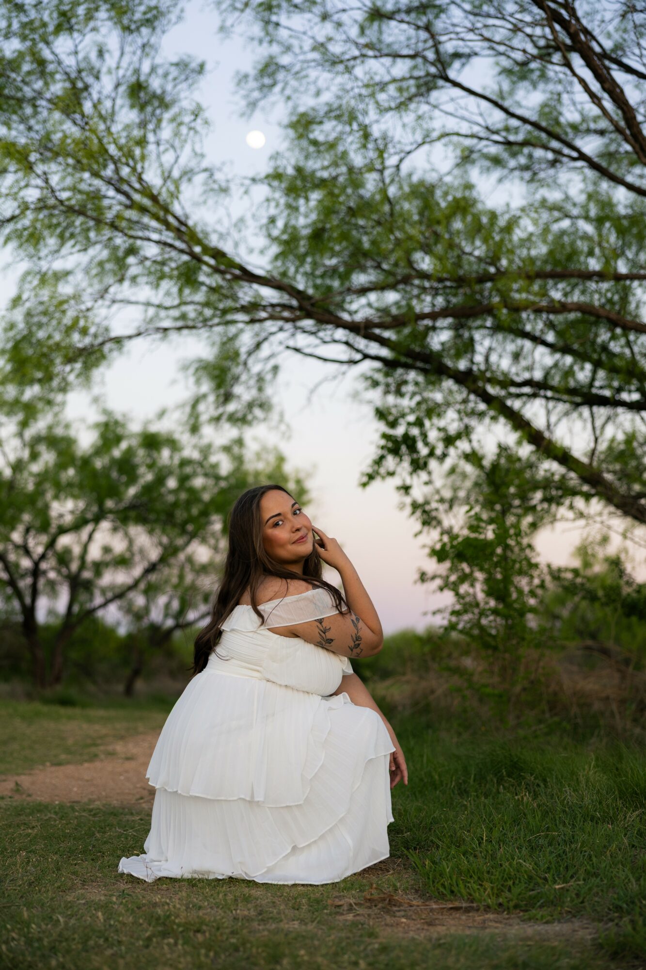 Woman in white dress sitting outdoors under trees, looking up, with sky and moon visible above.