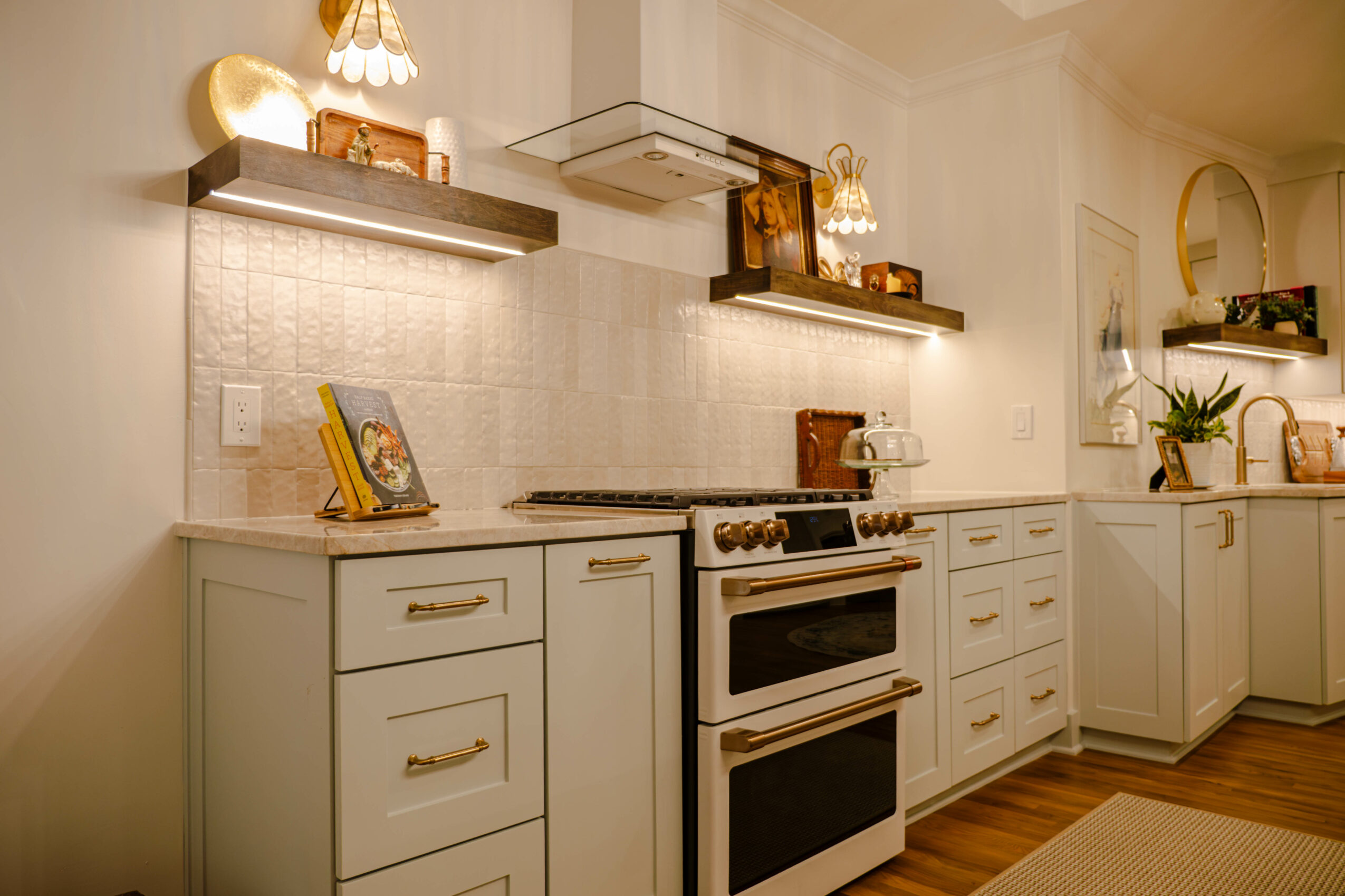Kitchen with white cabinets, stove, open shelves, decorative items, and potted plant, with wooden flooring and warm lighting.