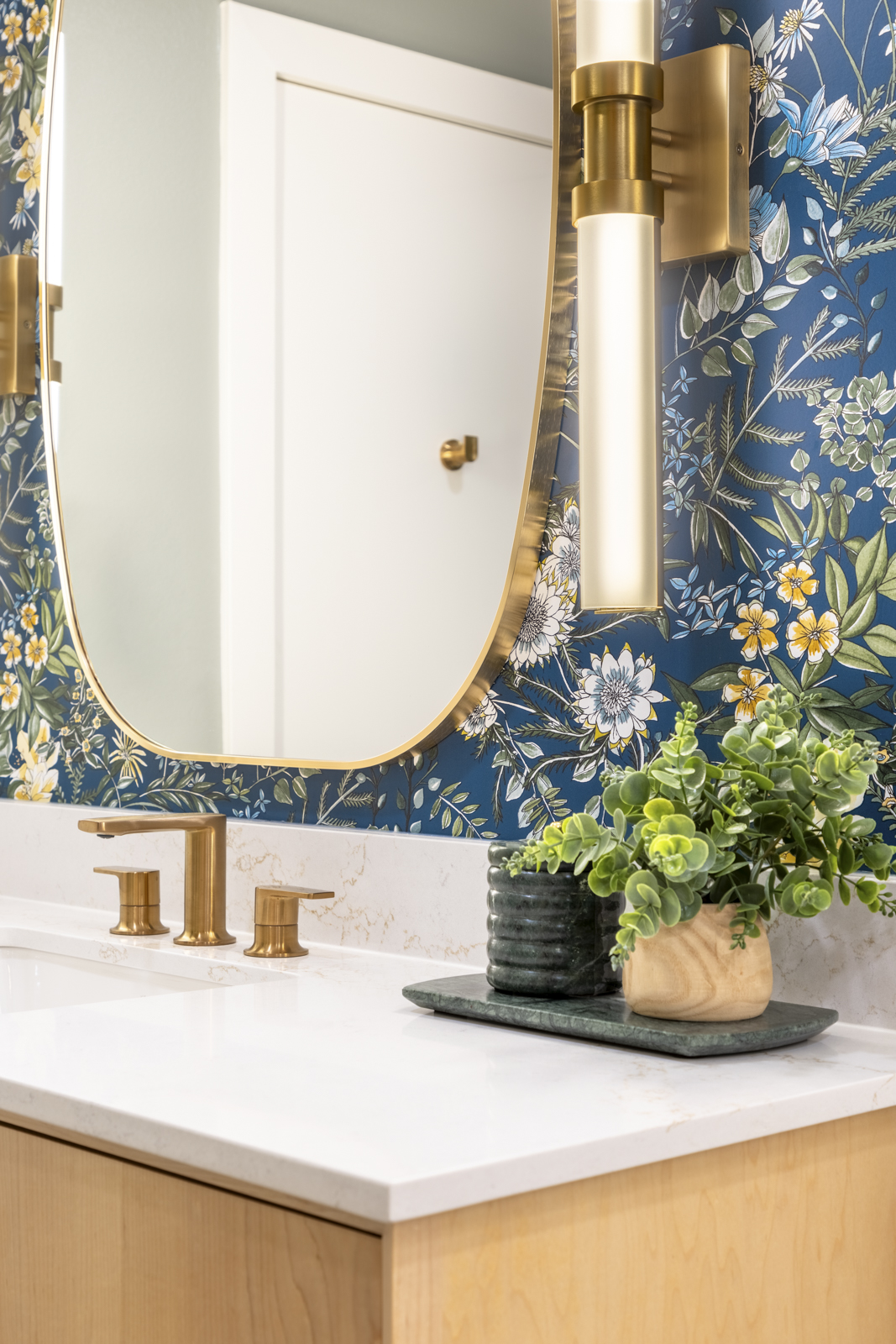 Bathroom mirror, floral wallpaper, wall-mounted light, plant in a pink pot, white countertop with gold fixtures.