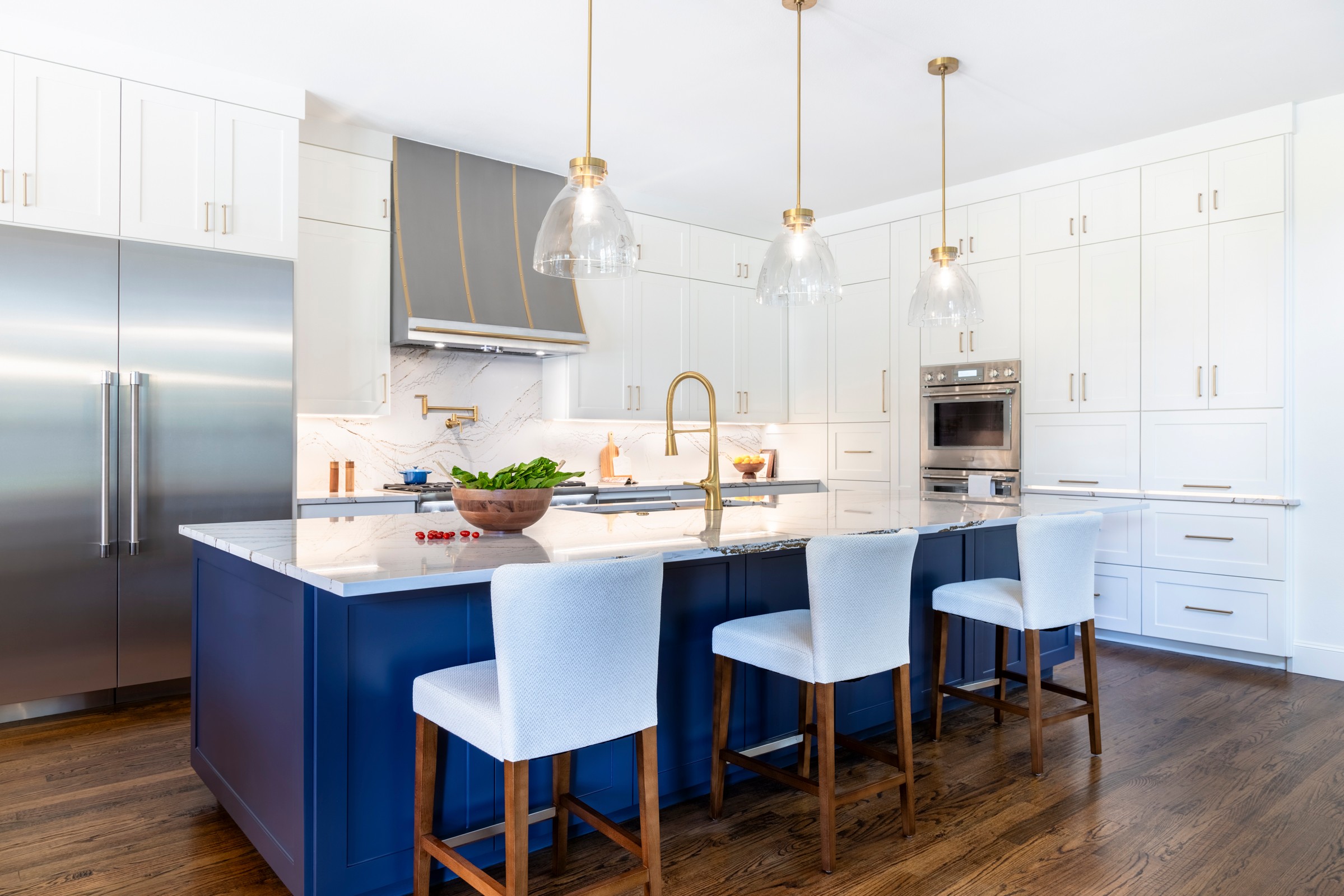 Modern kitchen with white cabinets, blue island, three white chairs, stainless steel appliances, and pendant lights.