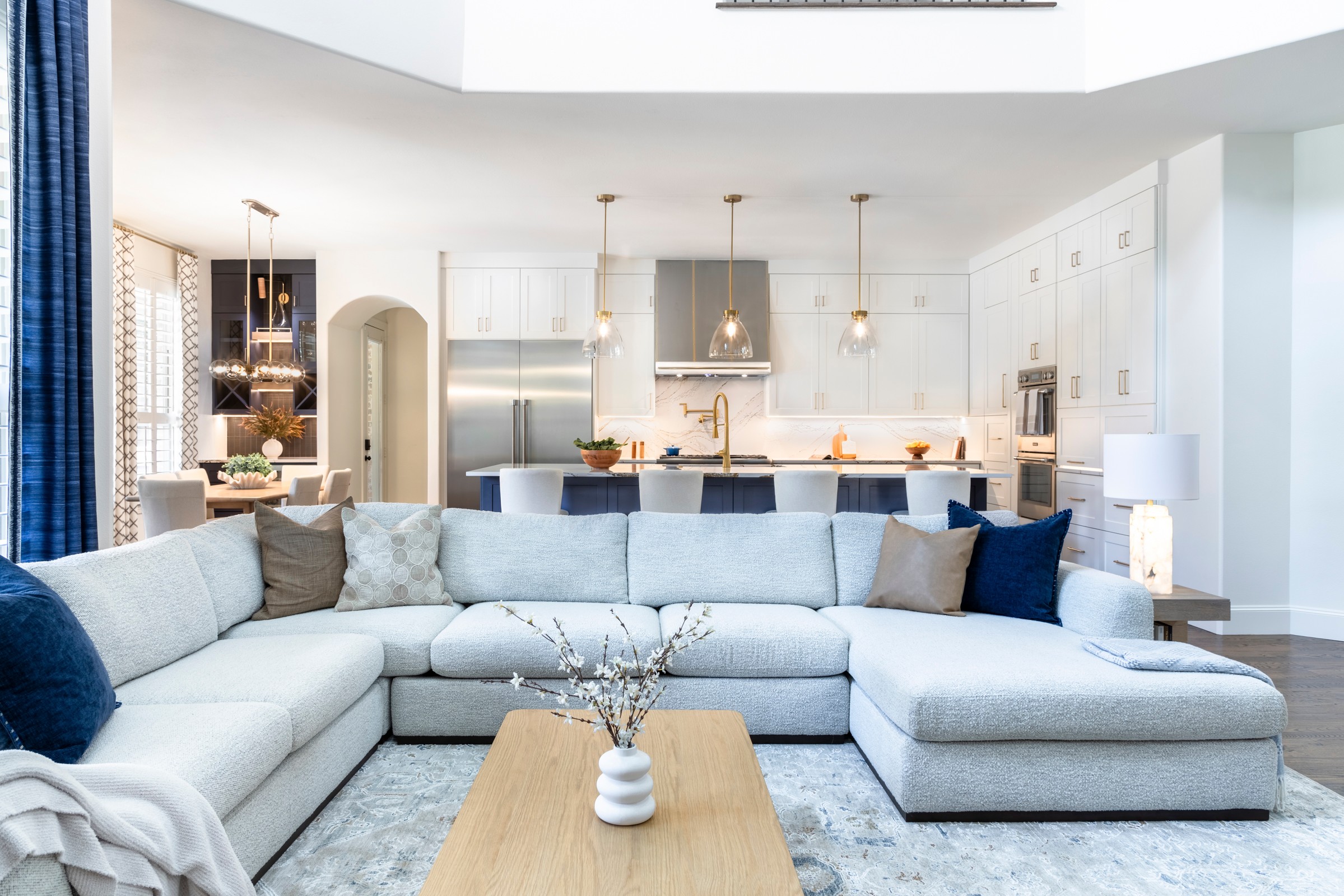 Open-concept living room with large sectional sofa, coffee table, and kitchen in background with white cabinets and pendant lights.