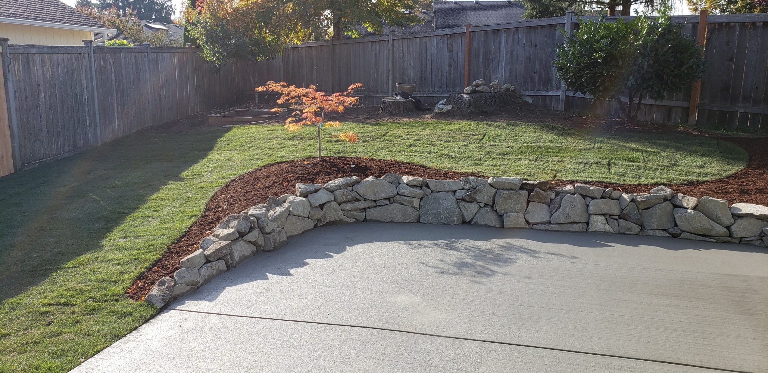 Yard with grass, stone border, wooden fence, and small trees, sunlight casting shadows.
