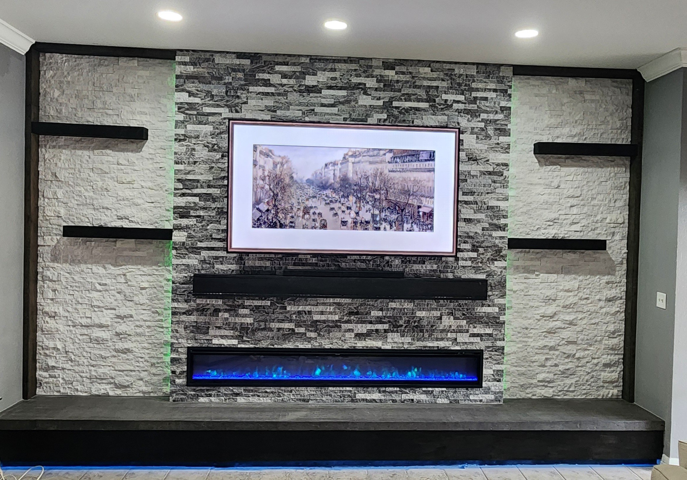 Wall with textured stone finish, mounted TV, and black shelves on each side, with a black entertainment unit below.