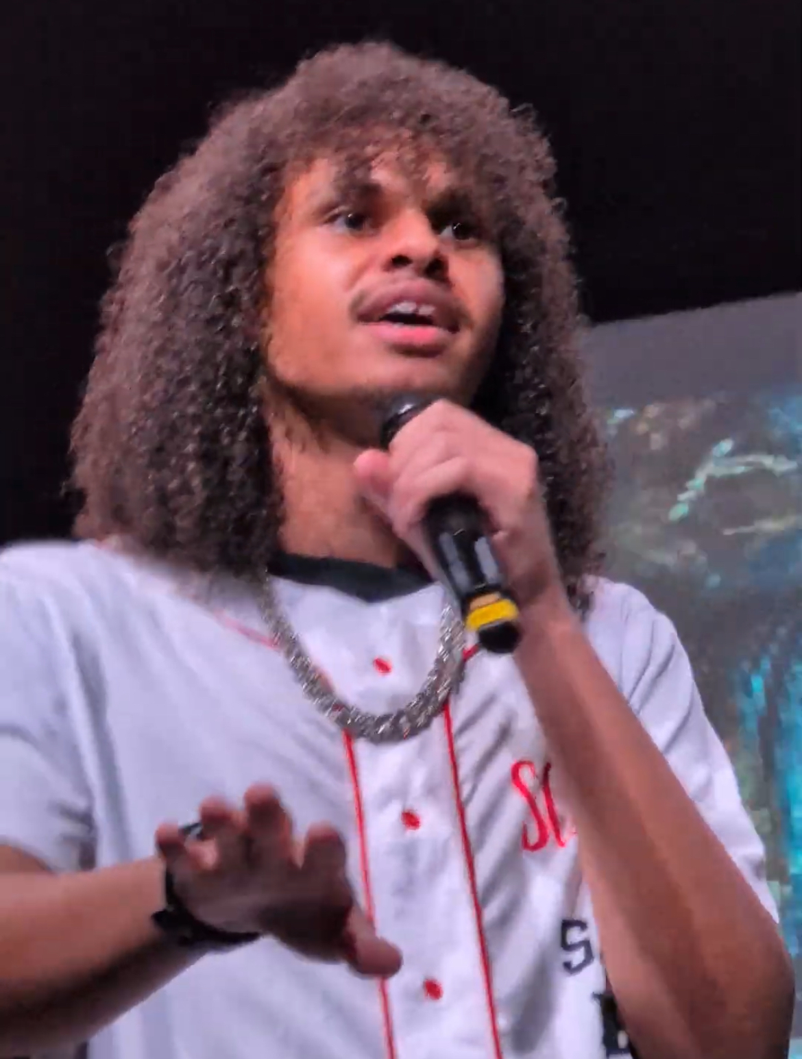 Person with curly hair holding a microphone, speaking or singing, wearing a light-colored shirt and a chain necklace.