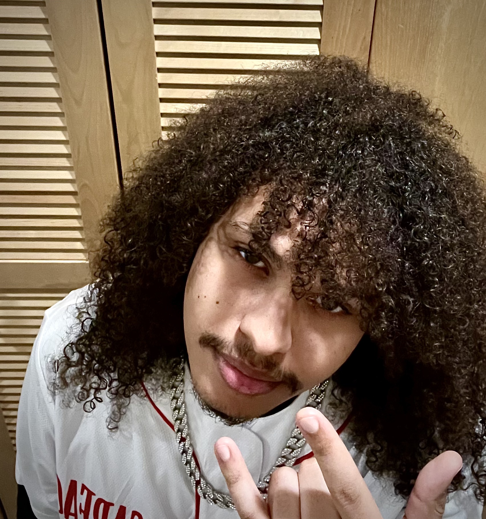 Person with curly hair and mustache making a hand gesture, wearing a white shirt with red text.
