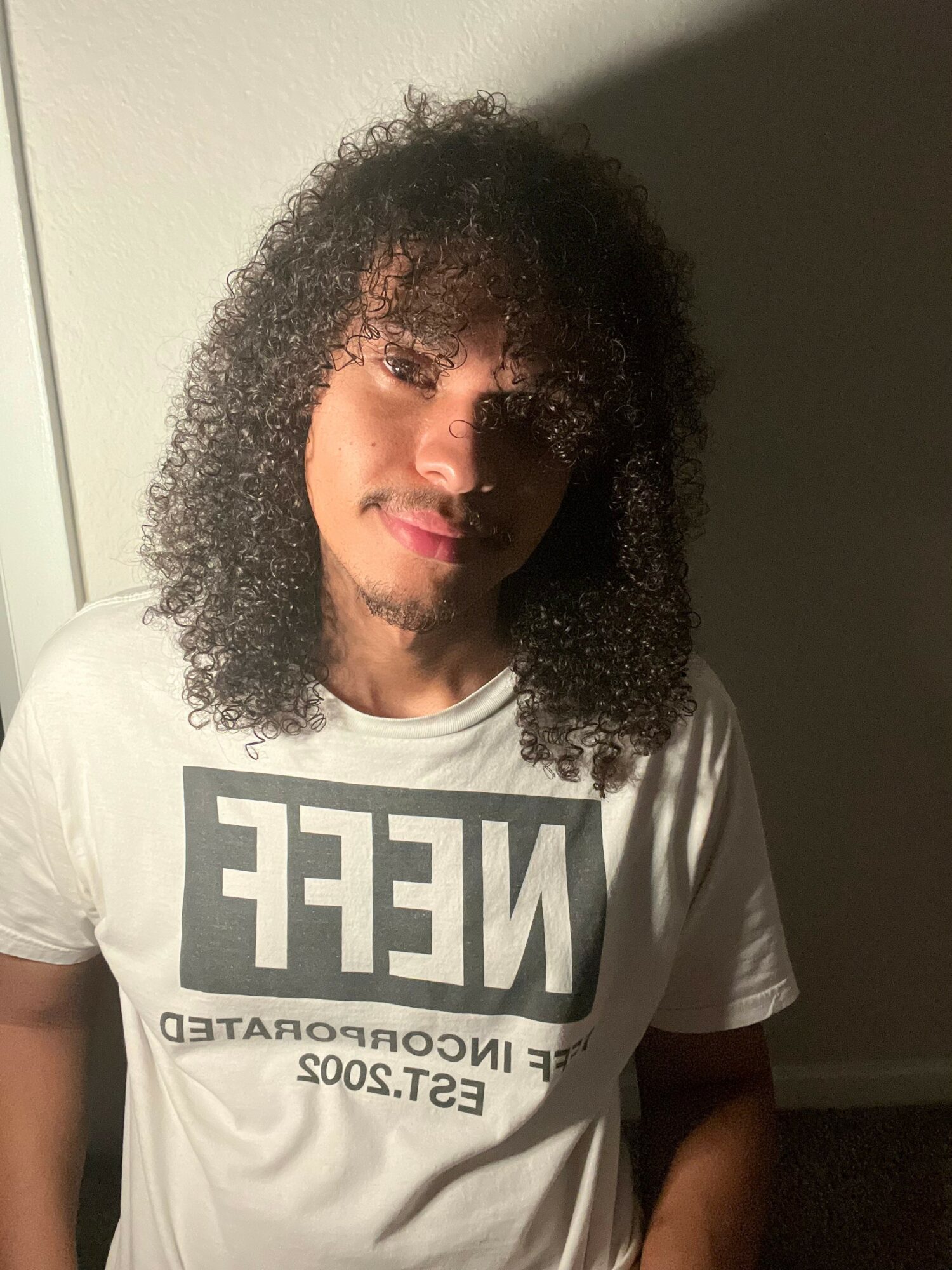 Young person with curly hair wearing a white T-shirt with black text, standing against a plain background.