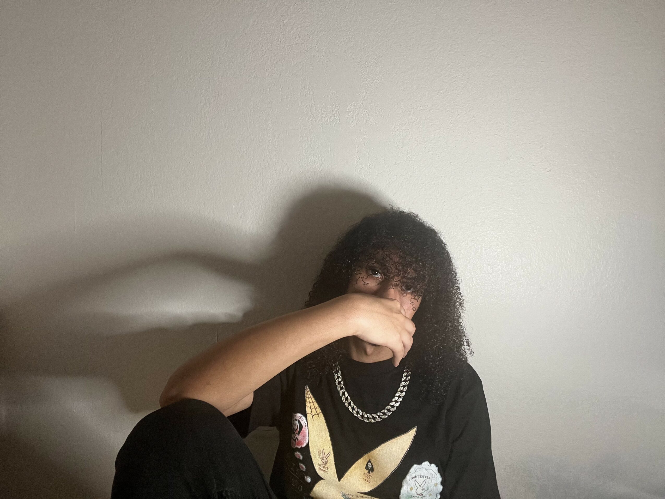 Person with curly hair covering face, wearing a black shirt with a chain necklace, against a plain wall.