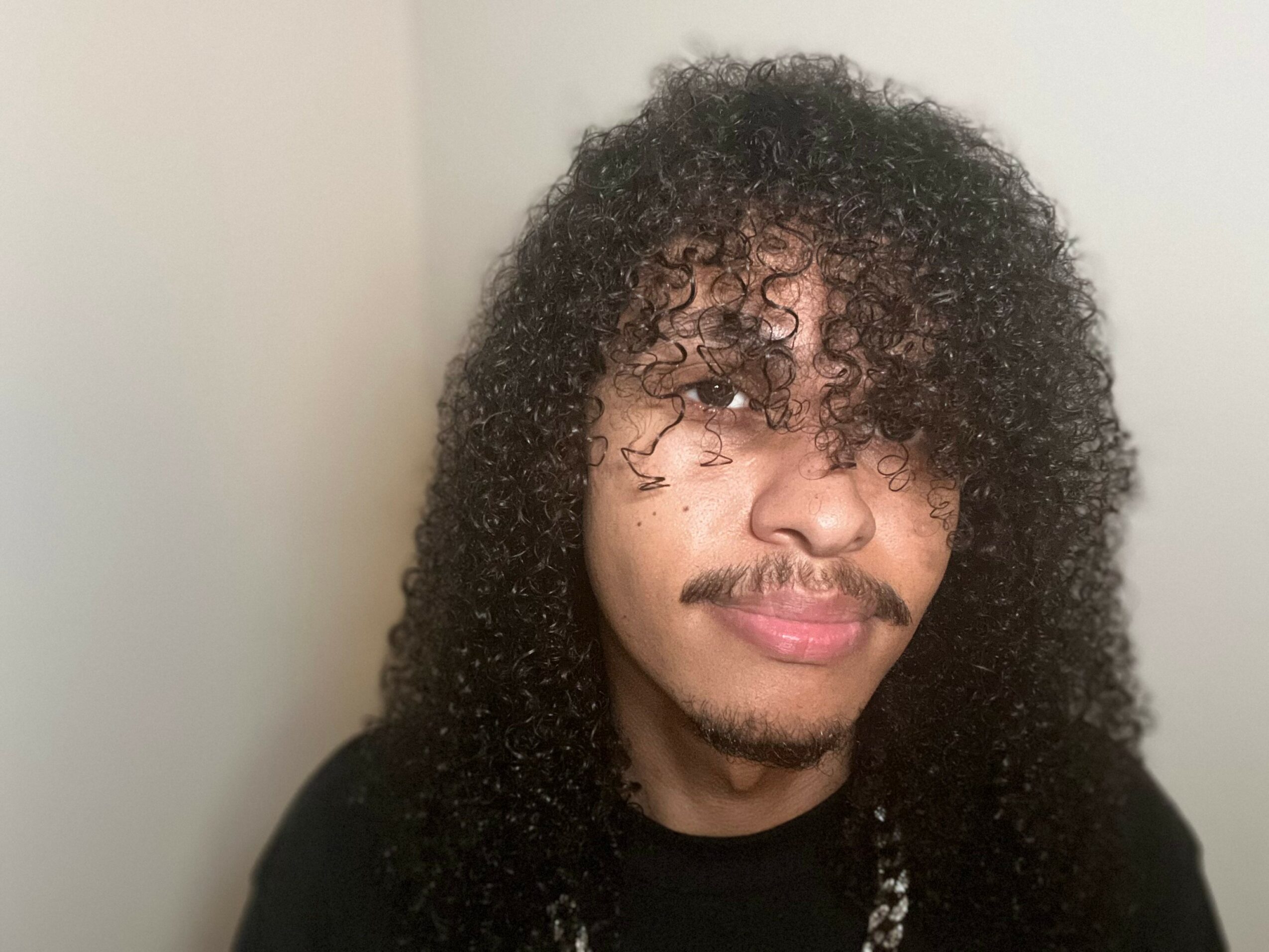 Young man with curly hair and mustache, smiling slightly, wearing a black shirt, against a plain background.