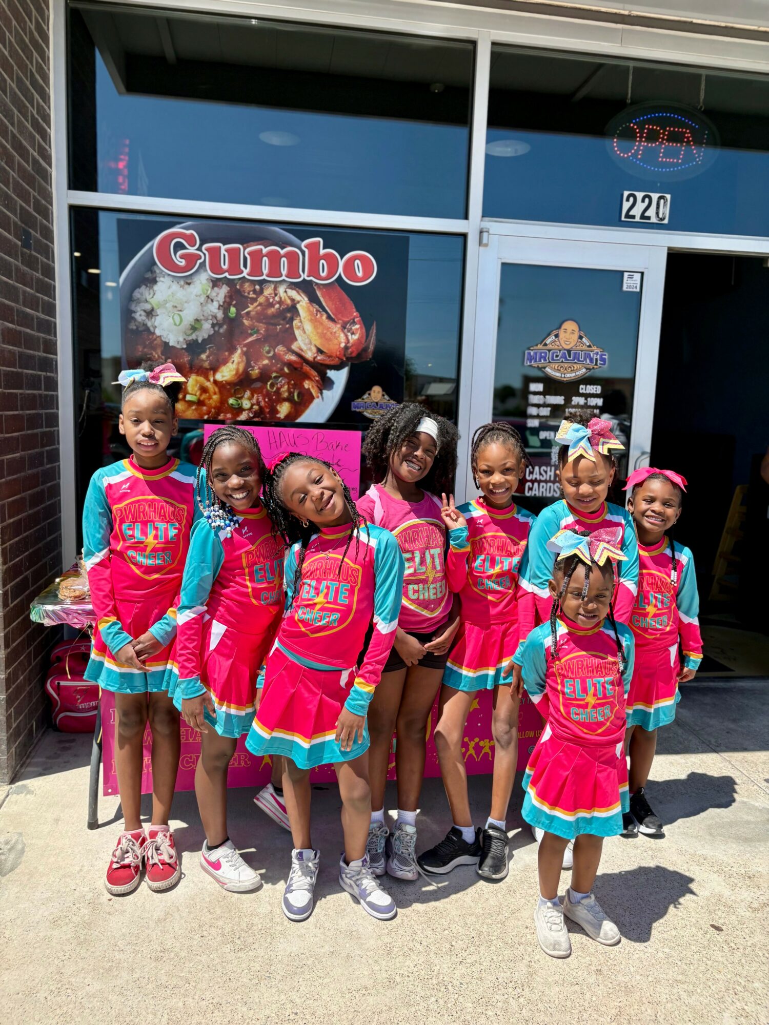 Group of seven children standing outside a store with a Gumbo sign, smiling and posing for the photo.