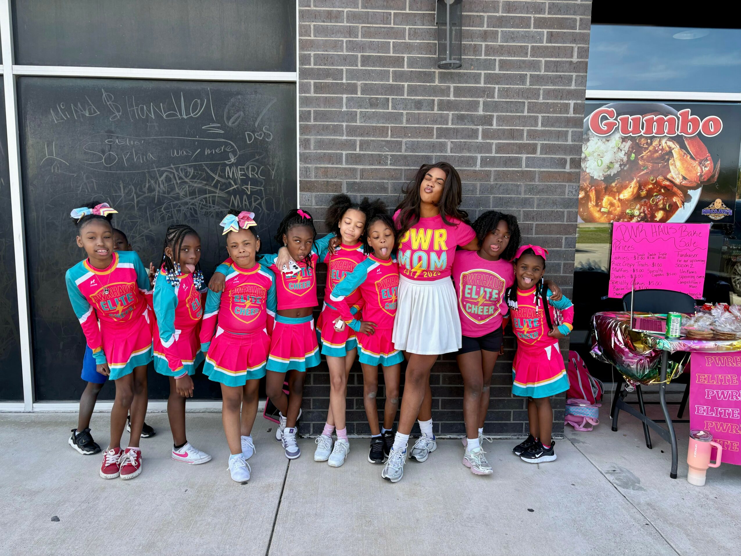 Group of young girls in pink and blue cheerleading outfits with a woman in a white dress, standing outside a building.