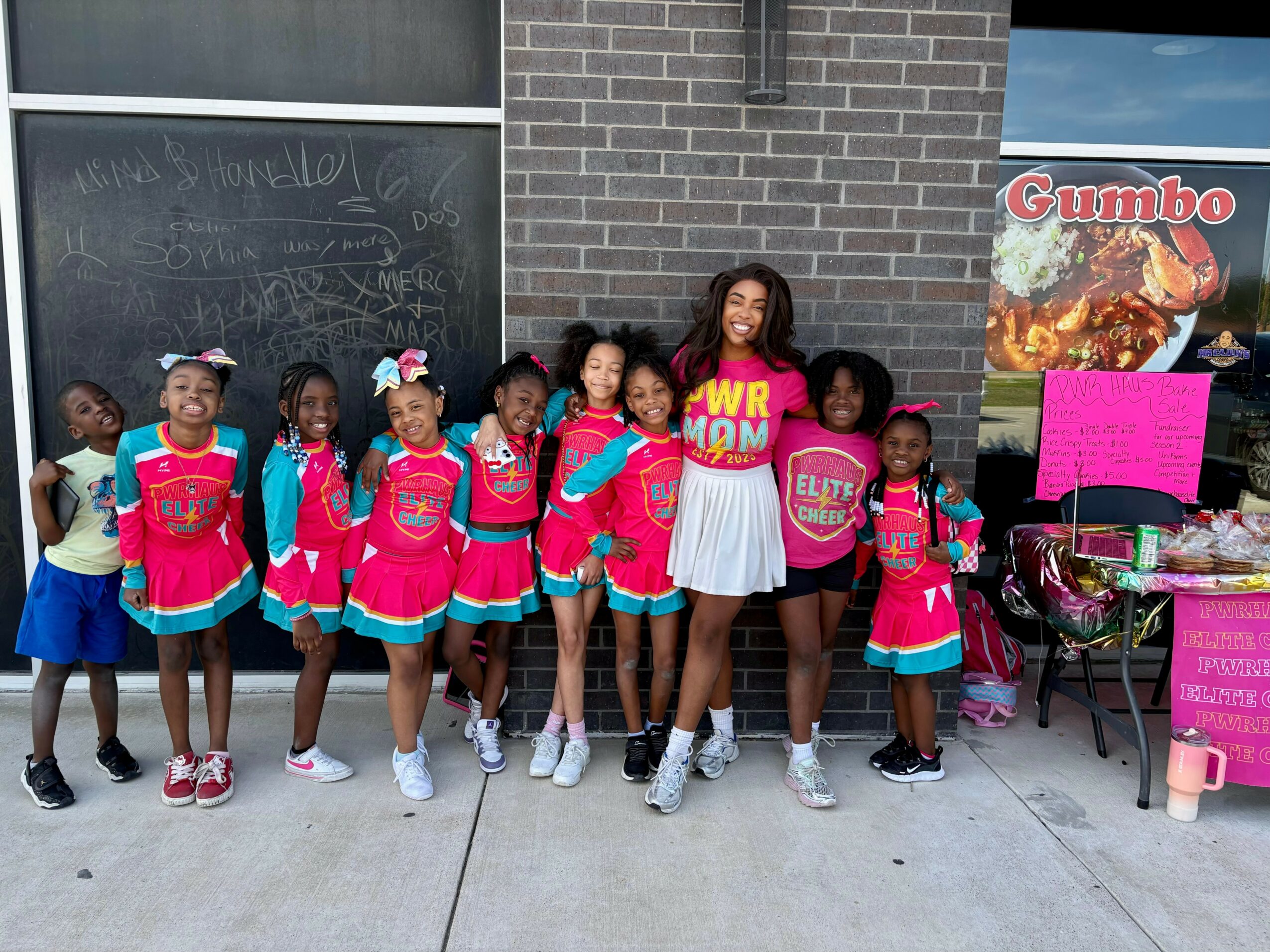Group of children and an adult standing outside in front of a brick wall, some wearing pink jackets and white skirts.
