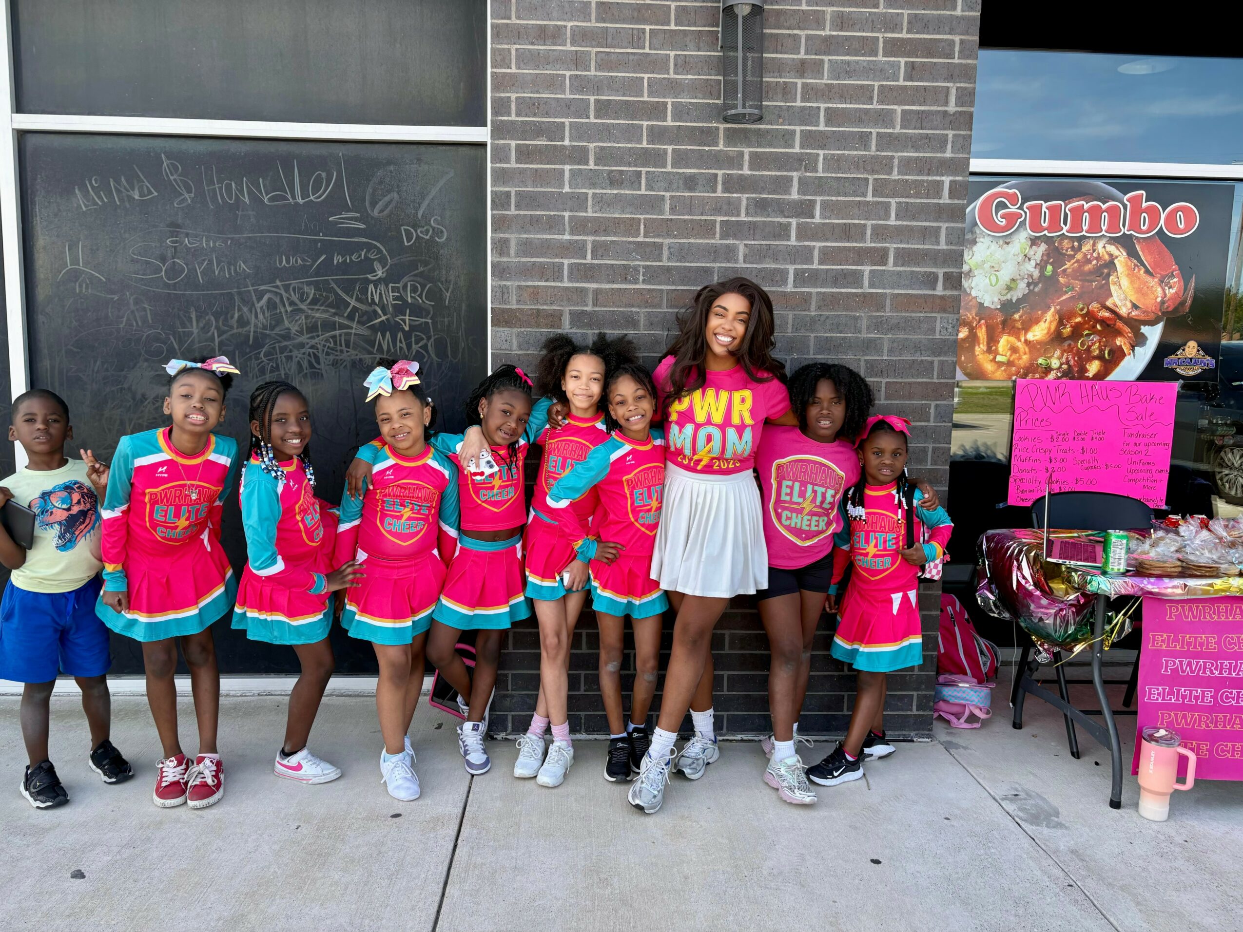 Group of young girls in pink and blue uniforms standing outside with woman in white dress, brick wall background.