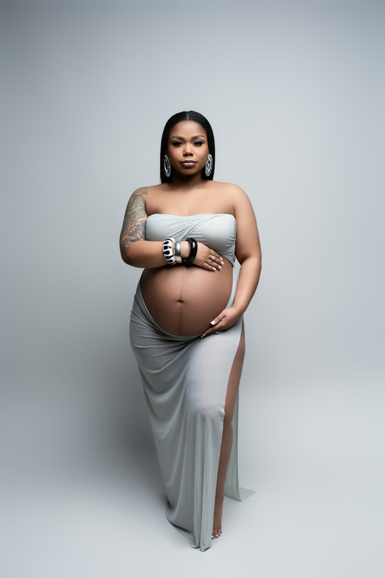 Pregnant woman in a strapless dress with tattoos, standing against a plain background, holding her belly with one hand.