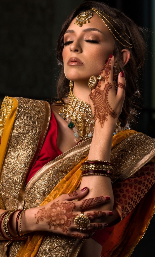 Woman in traditional attire with jewelry, eyes closed, hand near face, intricate henna on hands, ornate clothing.