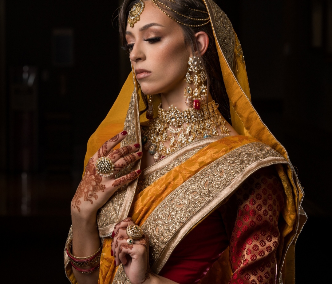 Woman dressed in traditional attire with jewelry, henna on hand, and a yellow head covering, looking down thoughtfully.