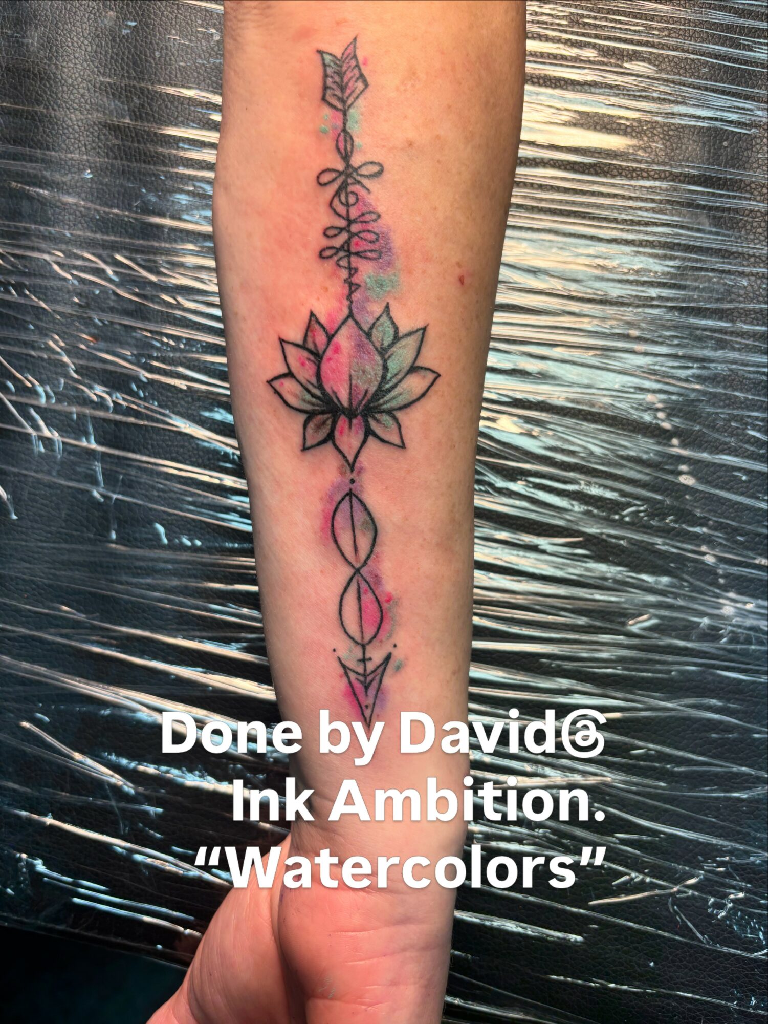 Vertical tattoo of a lotus flower with an arrow and decorative elements on forearm.