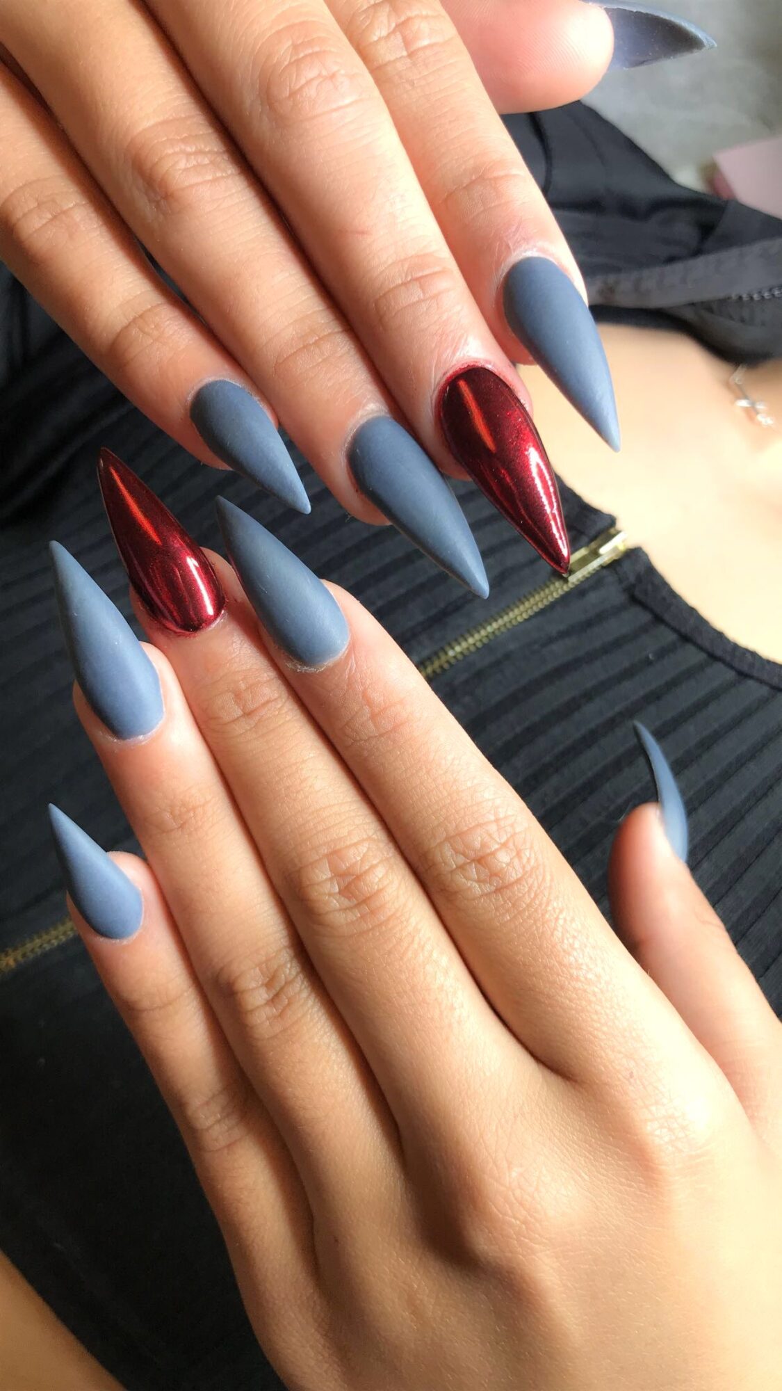 Hands with long, pointed nails painted in matte blue and shiny red colors, resting on a black fabric surface.