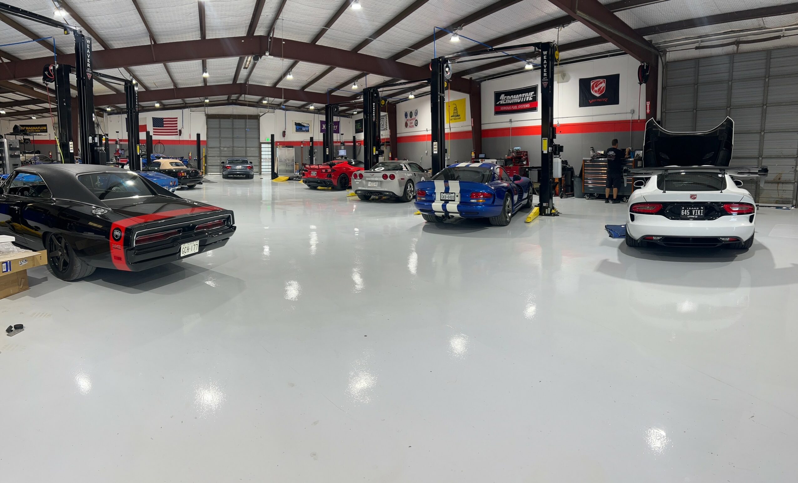 Indoor car workshop with several cars parked and workstations, high ceiling, and bright lighting.