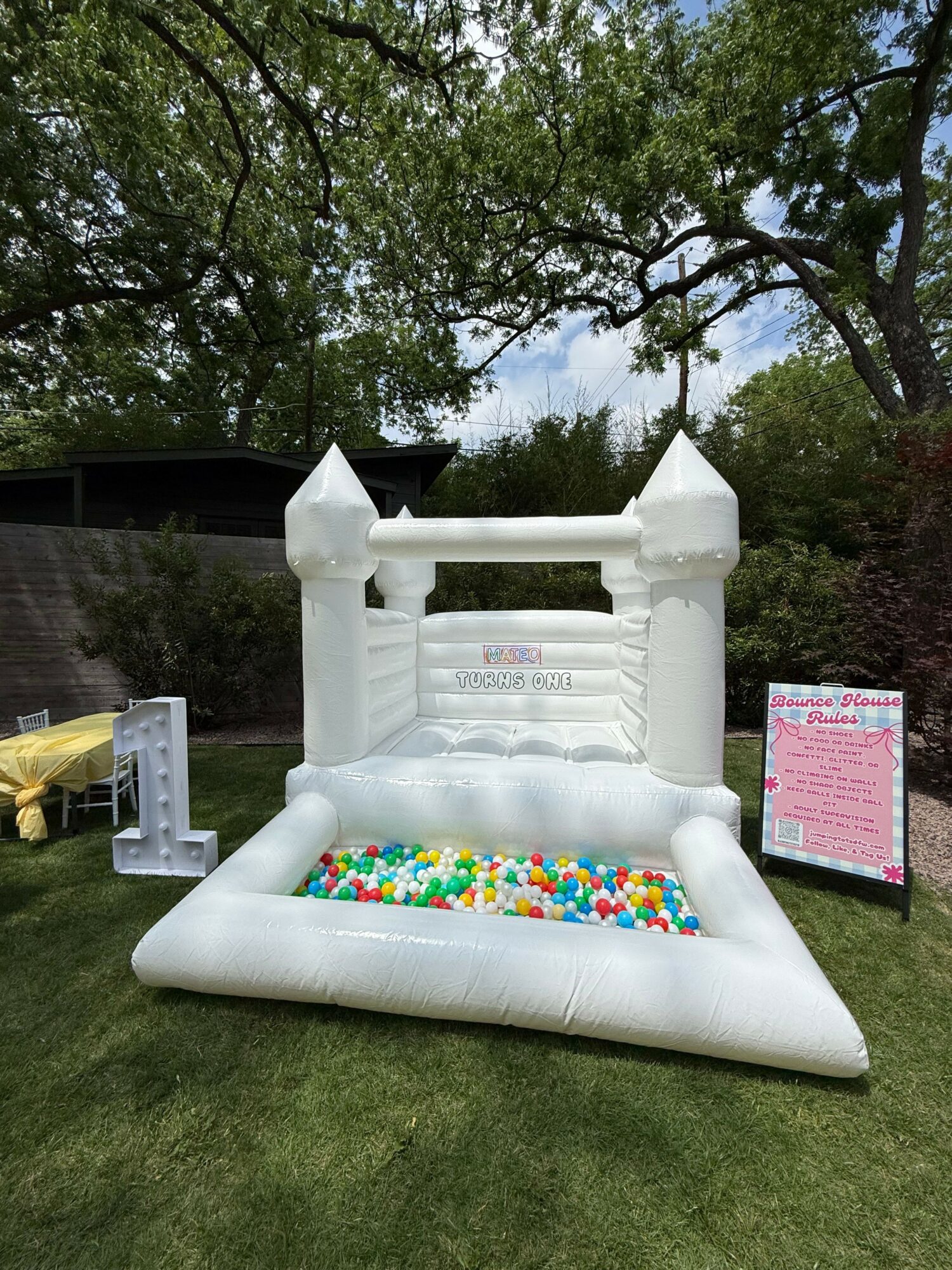 Inflatable castle bounce house with colorful balls inside, set outdoors on grass with trees and a sign nearby.