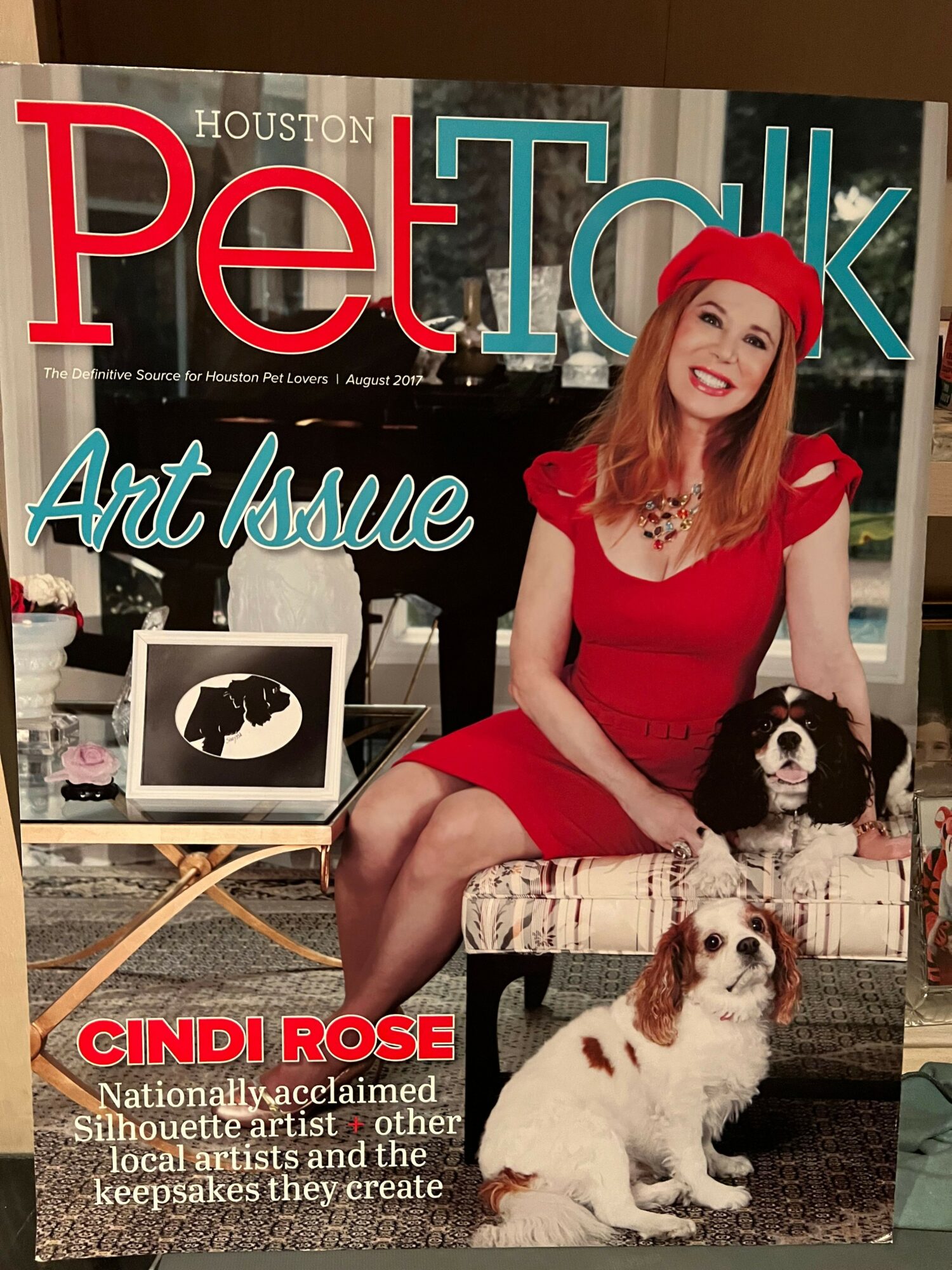 Woman in red dress sitting on a bench with two dogs, magazine cover with text and images in background.