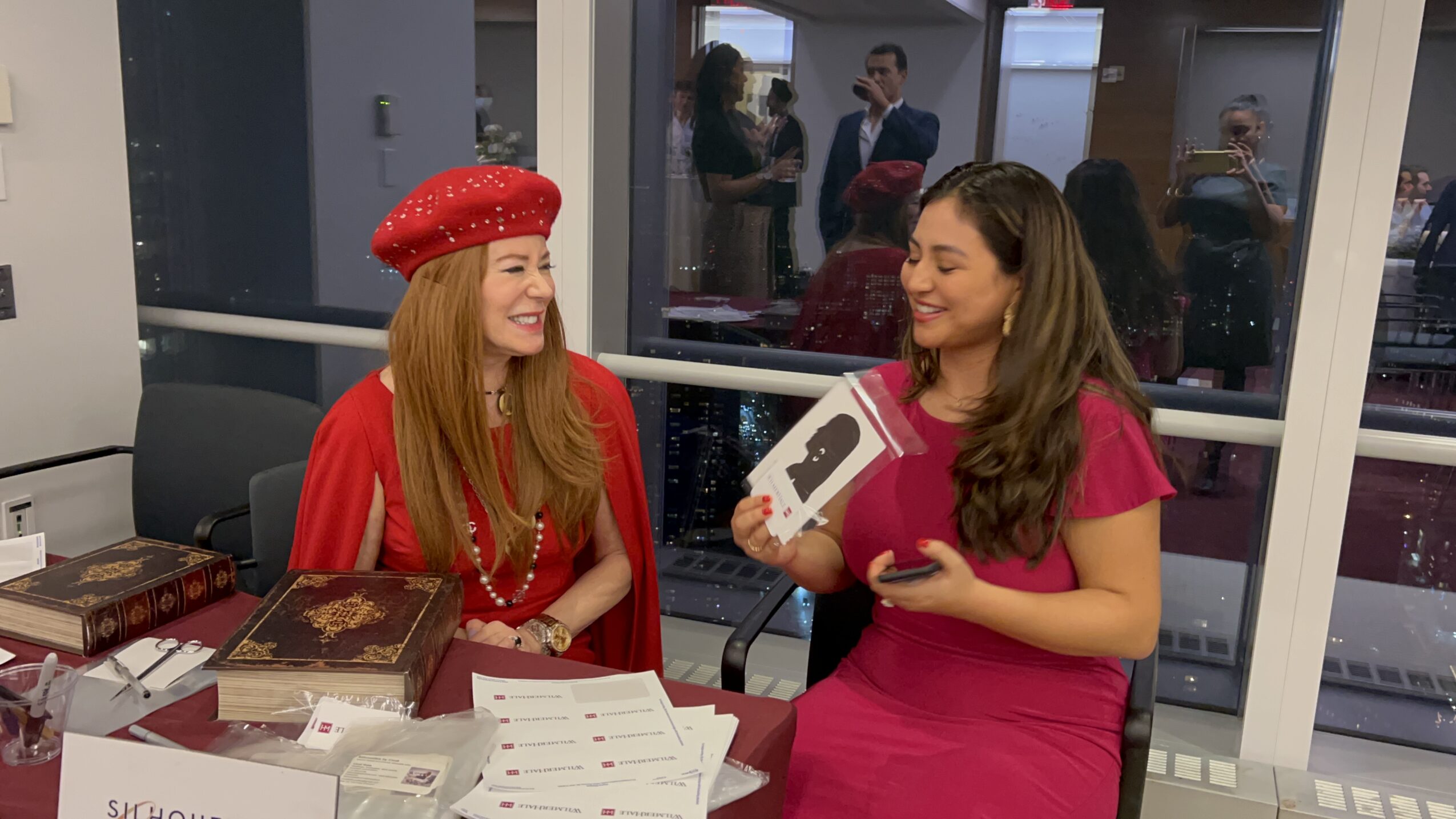 Two women sitting at a table, smiling and talking, with books and papers in front of them.