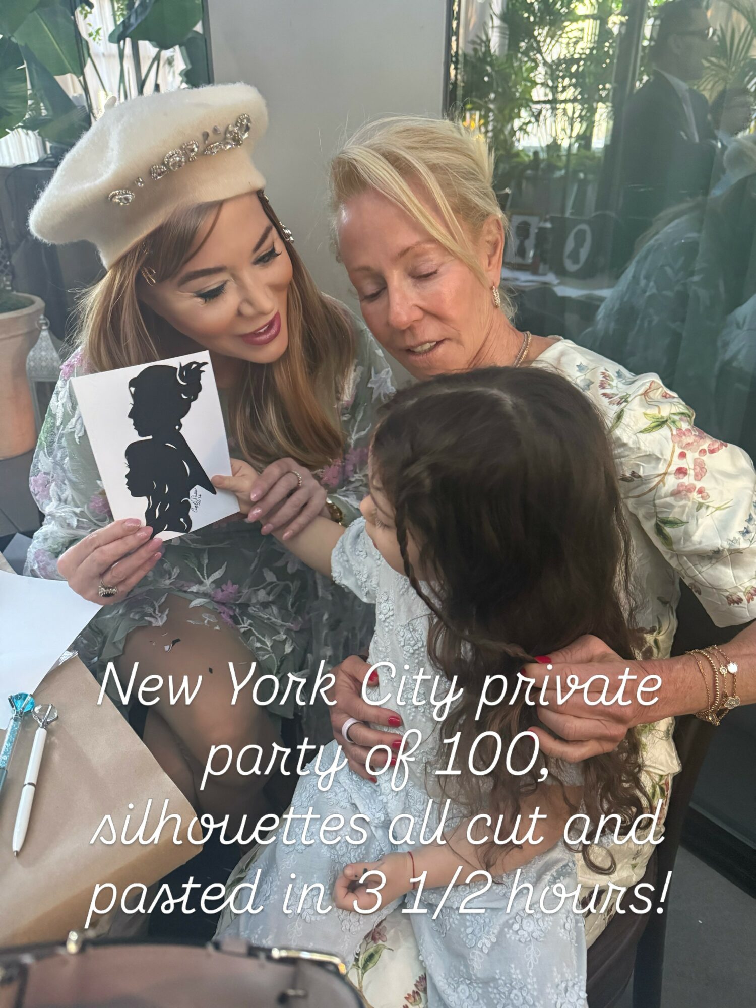 Three women sitting together, one holding a silhouette cutout, in a bright indoor setting with greenery outside.