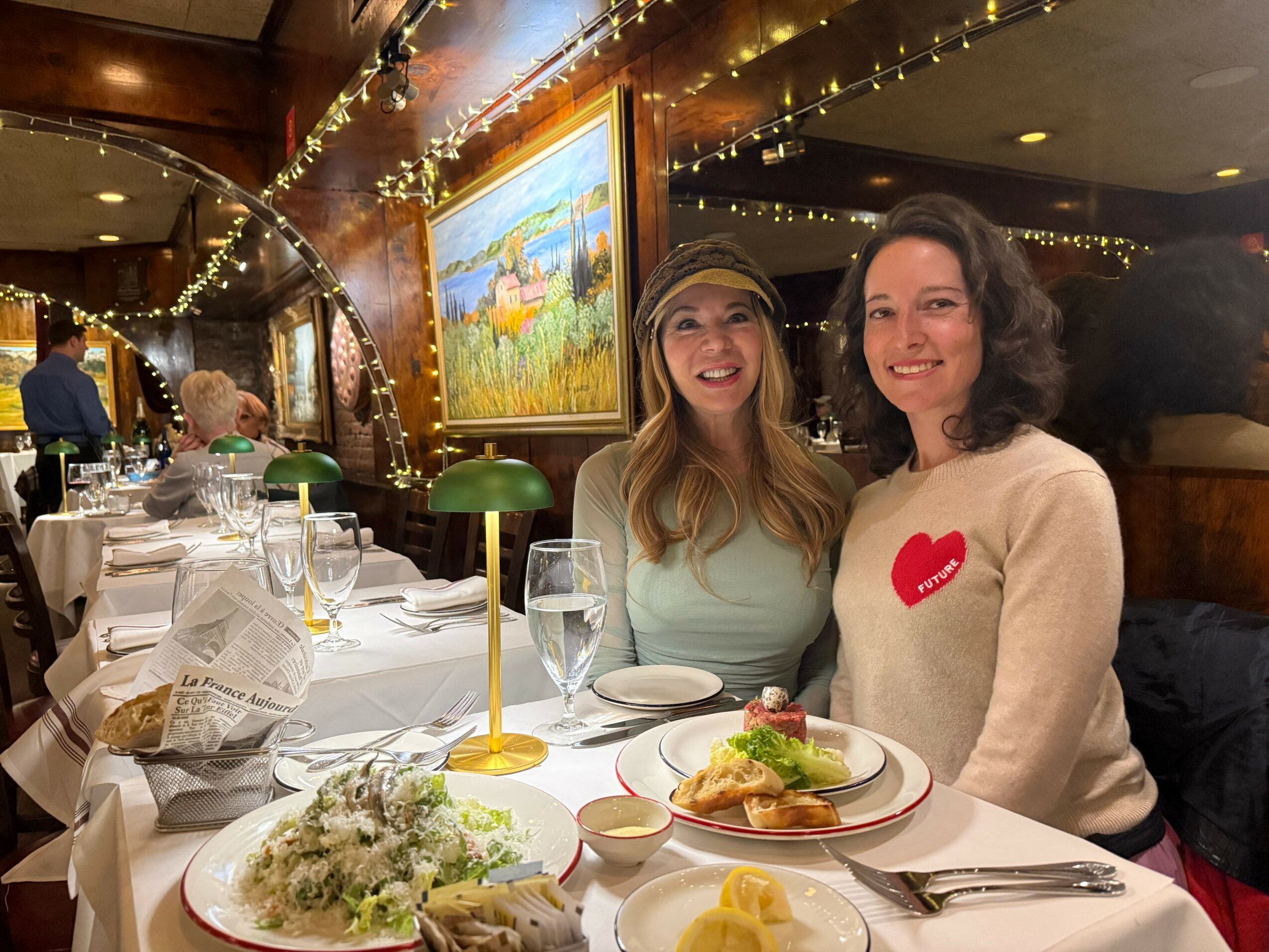 Two women smiling at a restaurant table with food and drinks, decorated with string lights and a painting in the background.