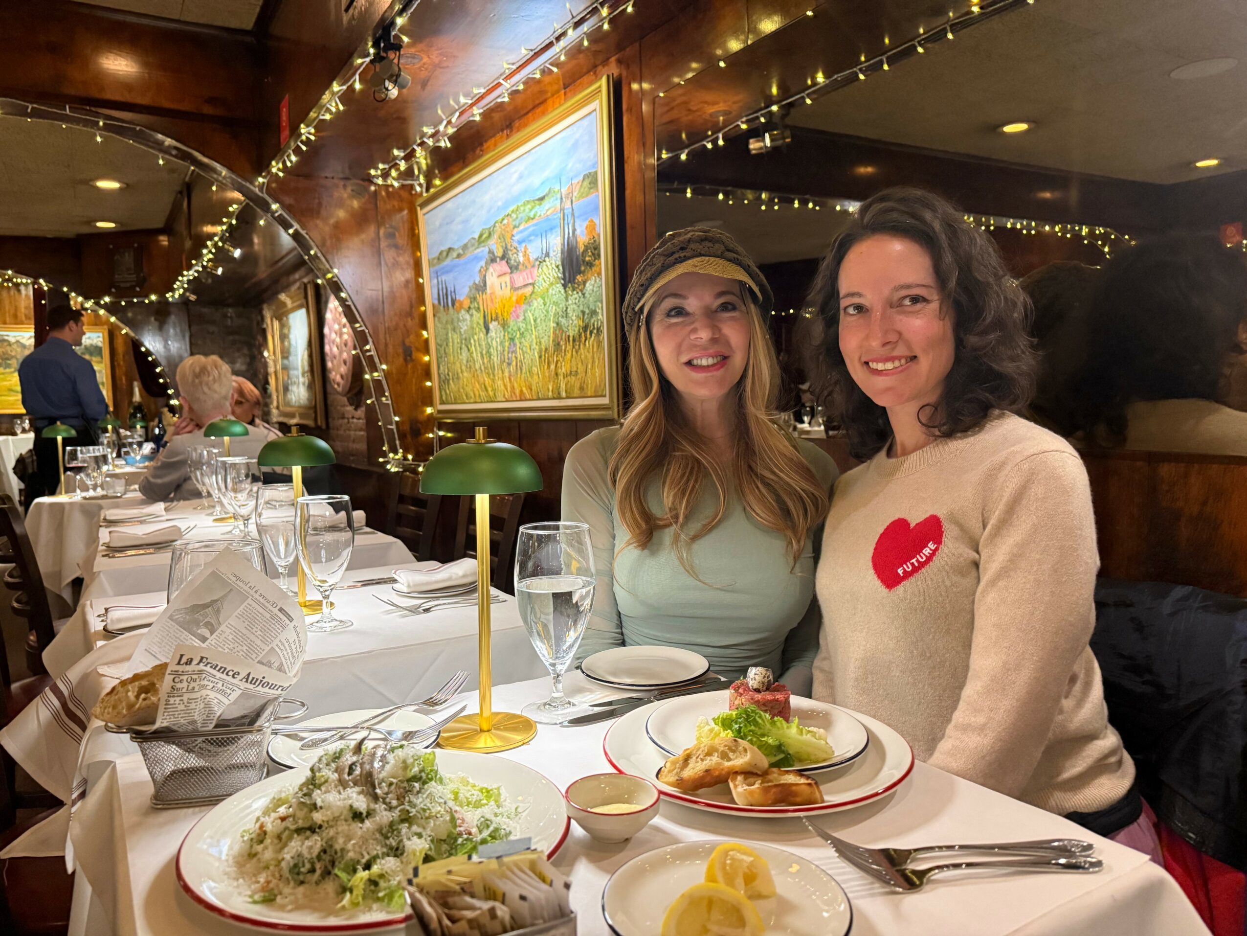 Two women smiling at a restaurant table with food, surrounded by decorated interior and other diners.